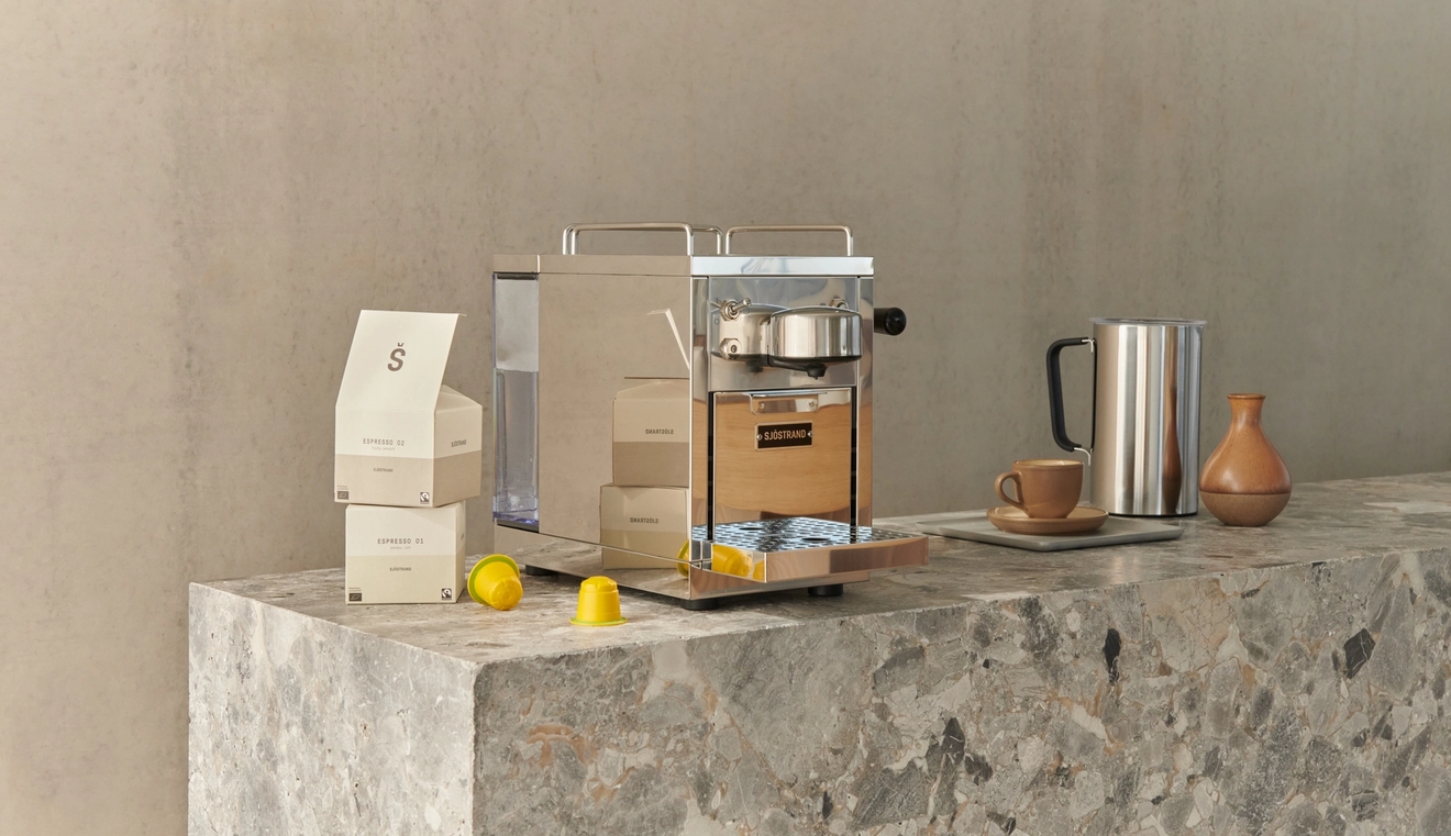 En beige og metallisk kapselkaffemaskin på en grå marmorbenk, med kaffeskuffer, gule kapsler, en metallkanne og en keramisk kopp.