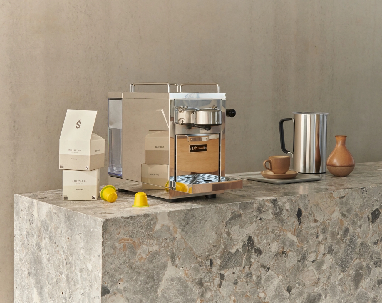 En beige og metallisk kapselkaffemaskin på en grå marmorbenk, med kaffeskuffer, gule kapsler, en metallkanne og en keramisk kopp.