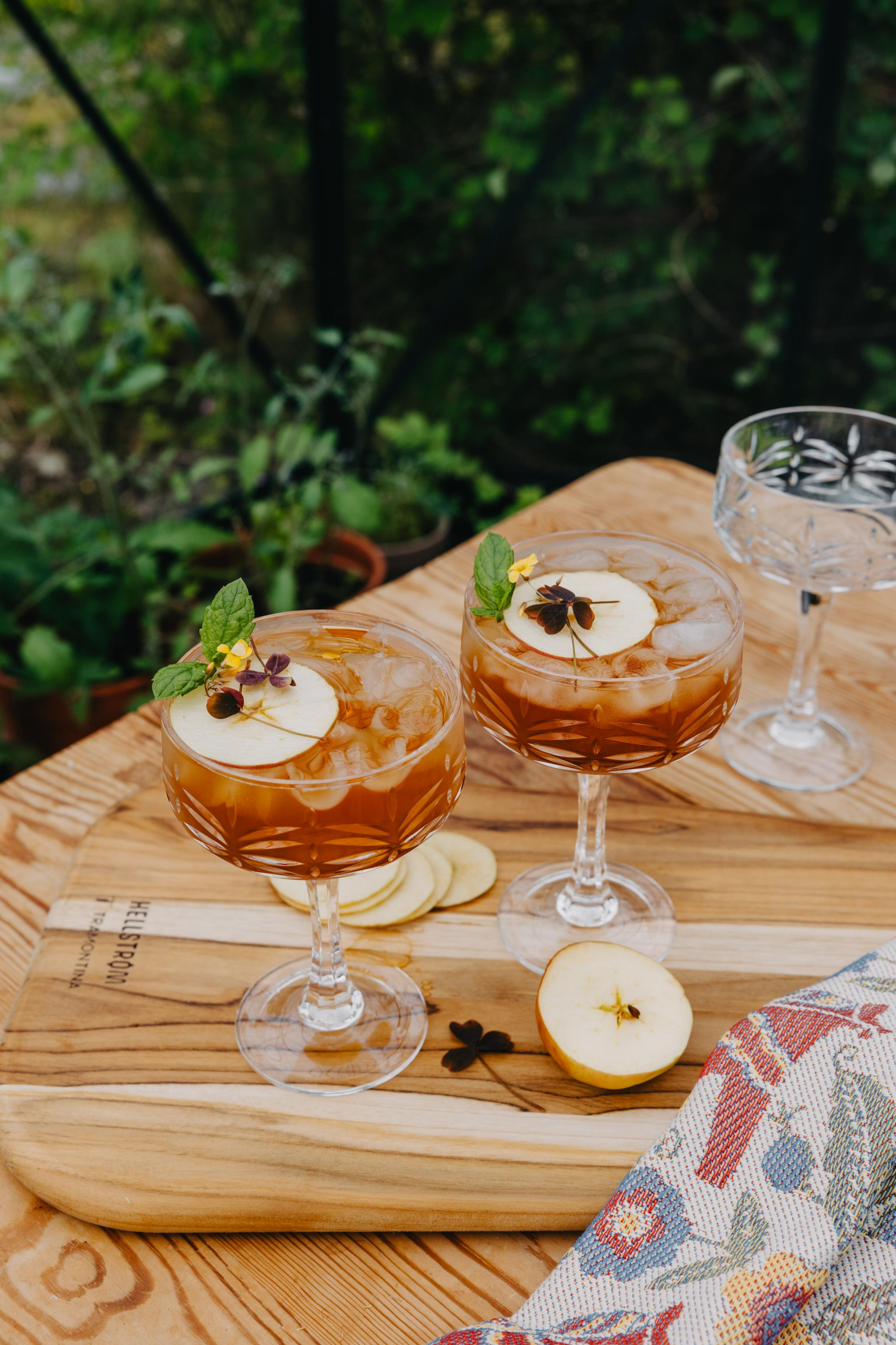 Två eleganta drinkar med äppelskivor och örter serveras i kristallglas på en träbricka utomhus.
