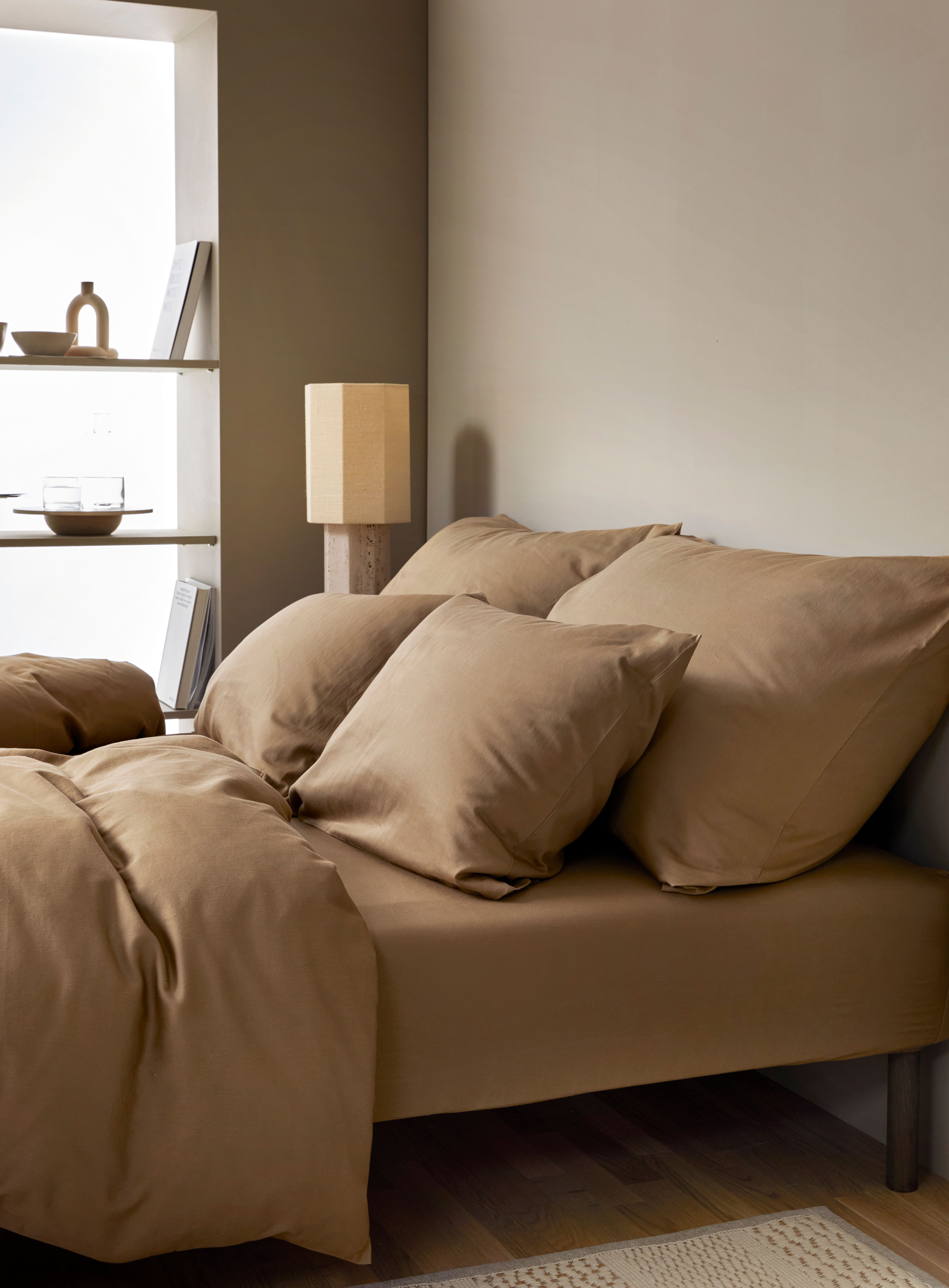 Sovrum med en stor säng bäddat i beige bruna sängkläder, i bakgrunden ett fönster med hyllplan