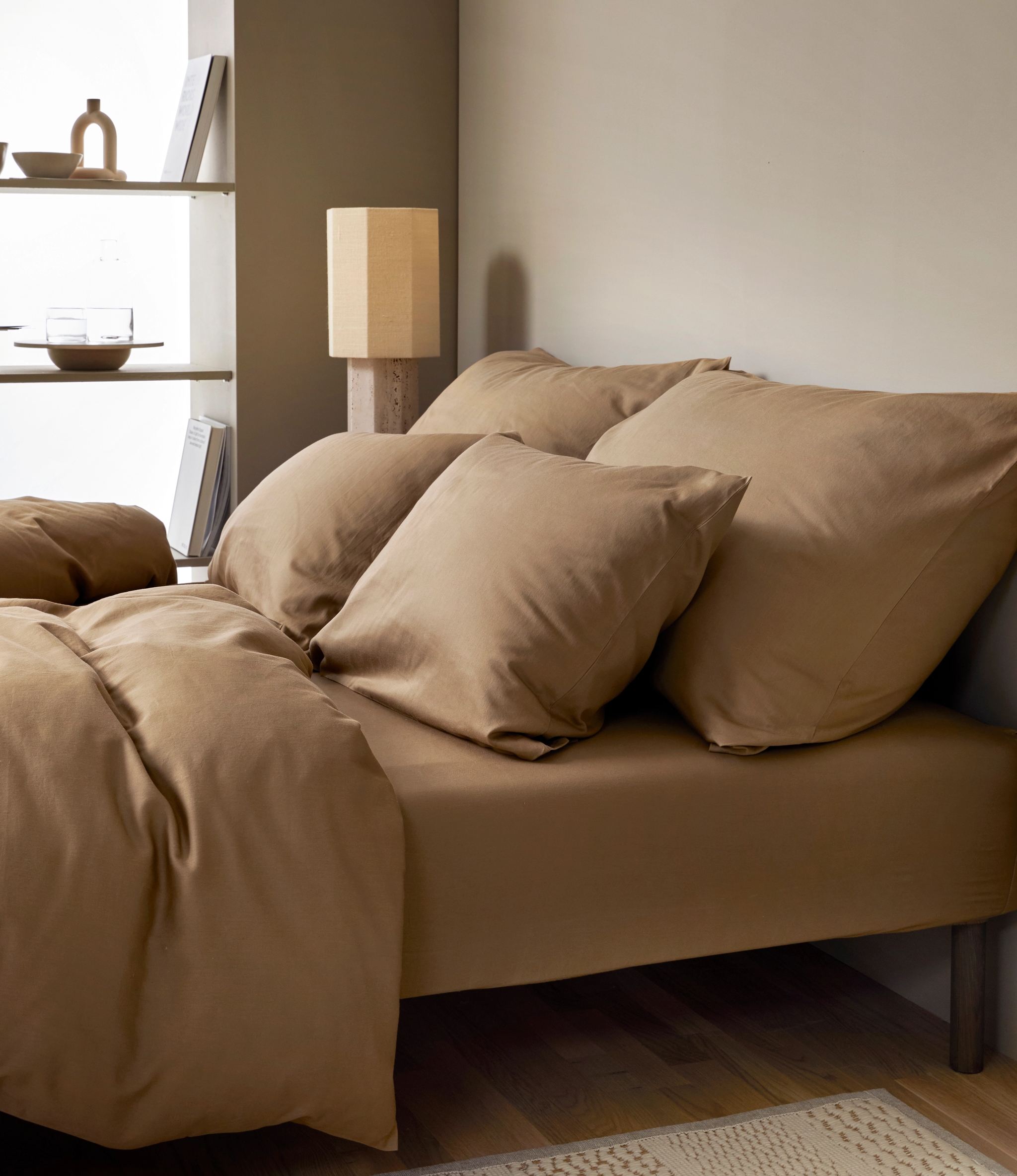 Sovrum med en stor säng bäddat i beige bruna sängkläder, i bakgrunden ett fönster med hyllplan