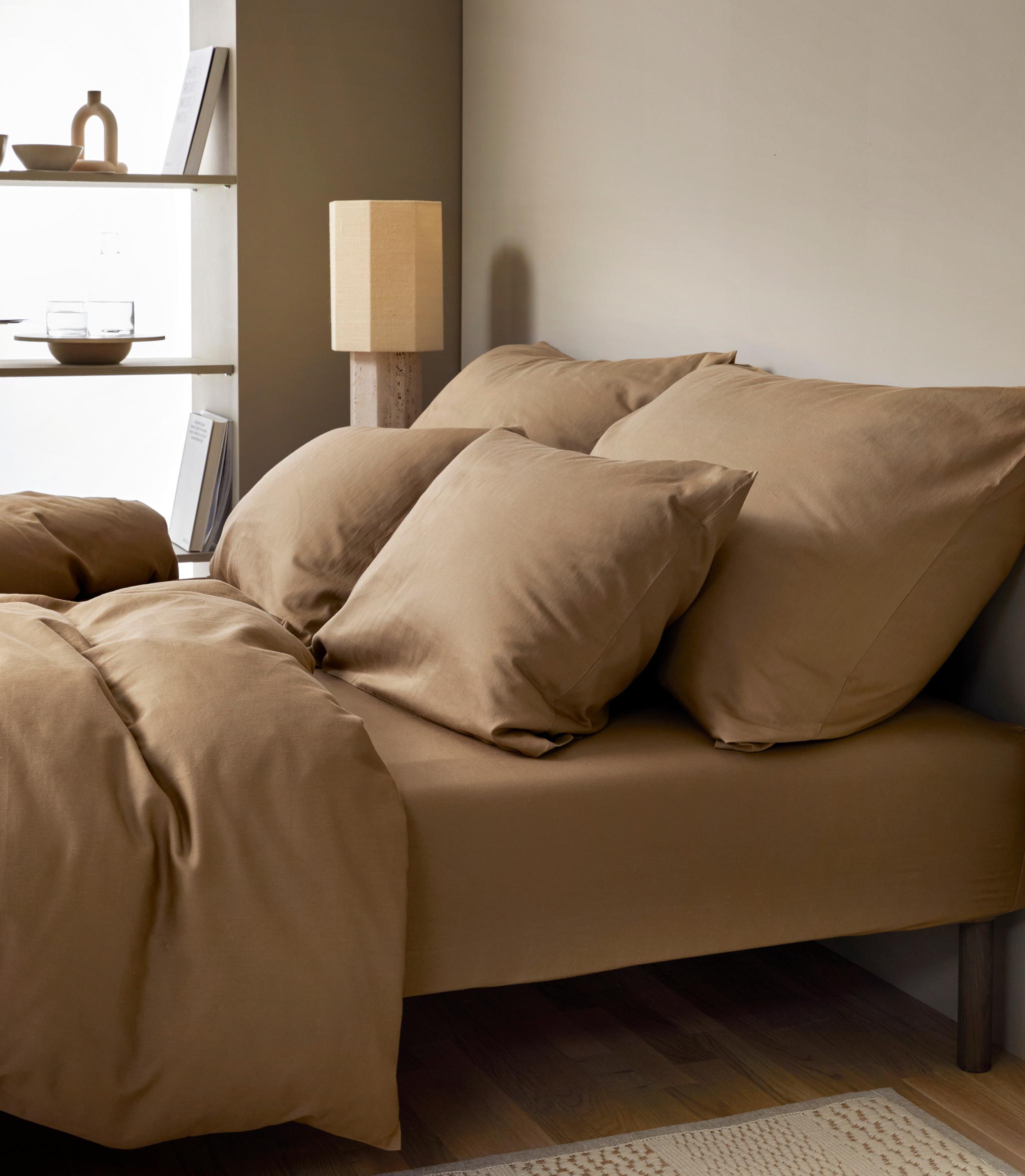 Sovrum med en stor säng bäddat i beige bruna sängkläder, i bakgrunden ett fönster med hyllplan