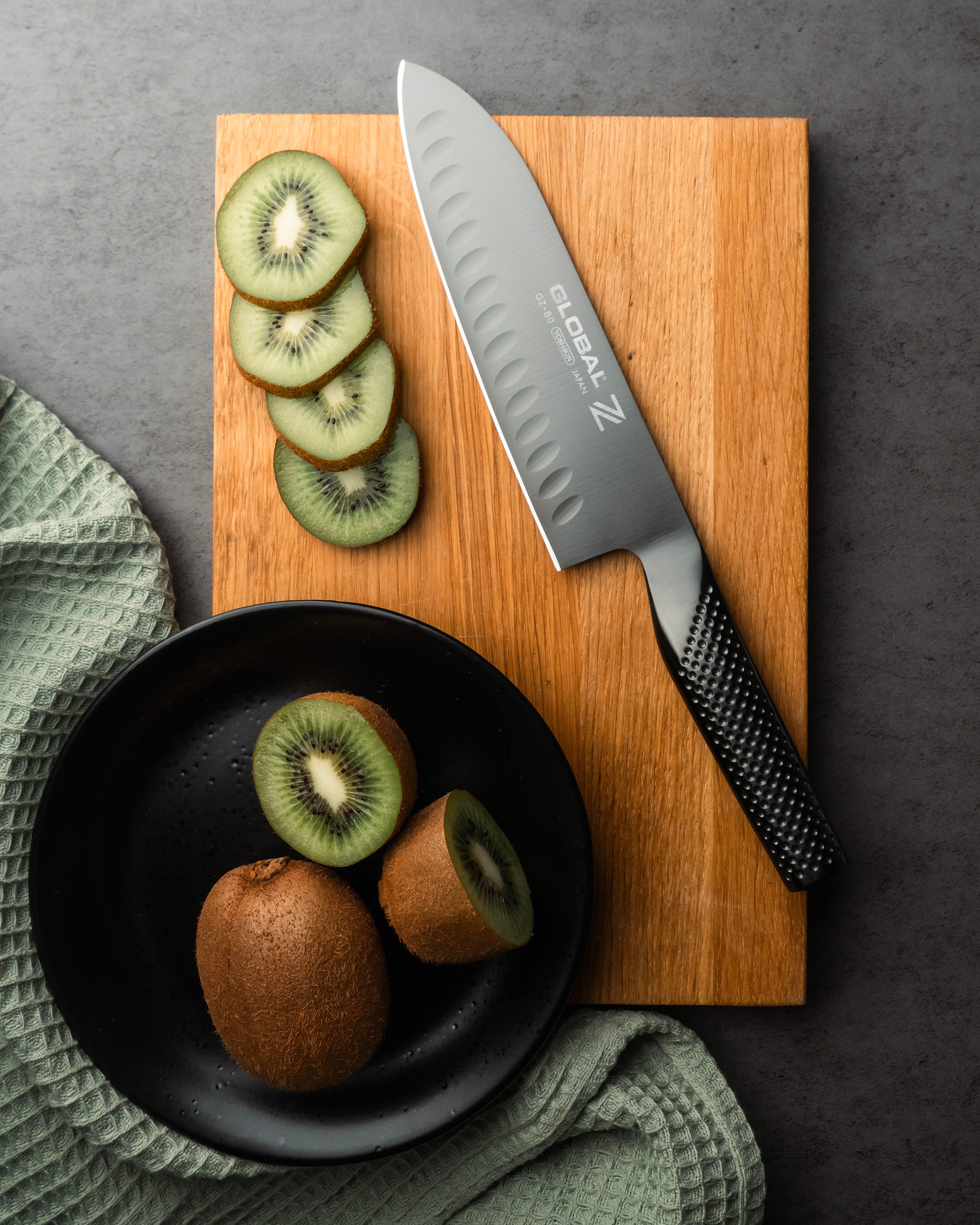 Skivad kiwi och en Global-kökskniv på en skärbräda bredvid en svart tallrik med färska kiwifrukter.