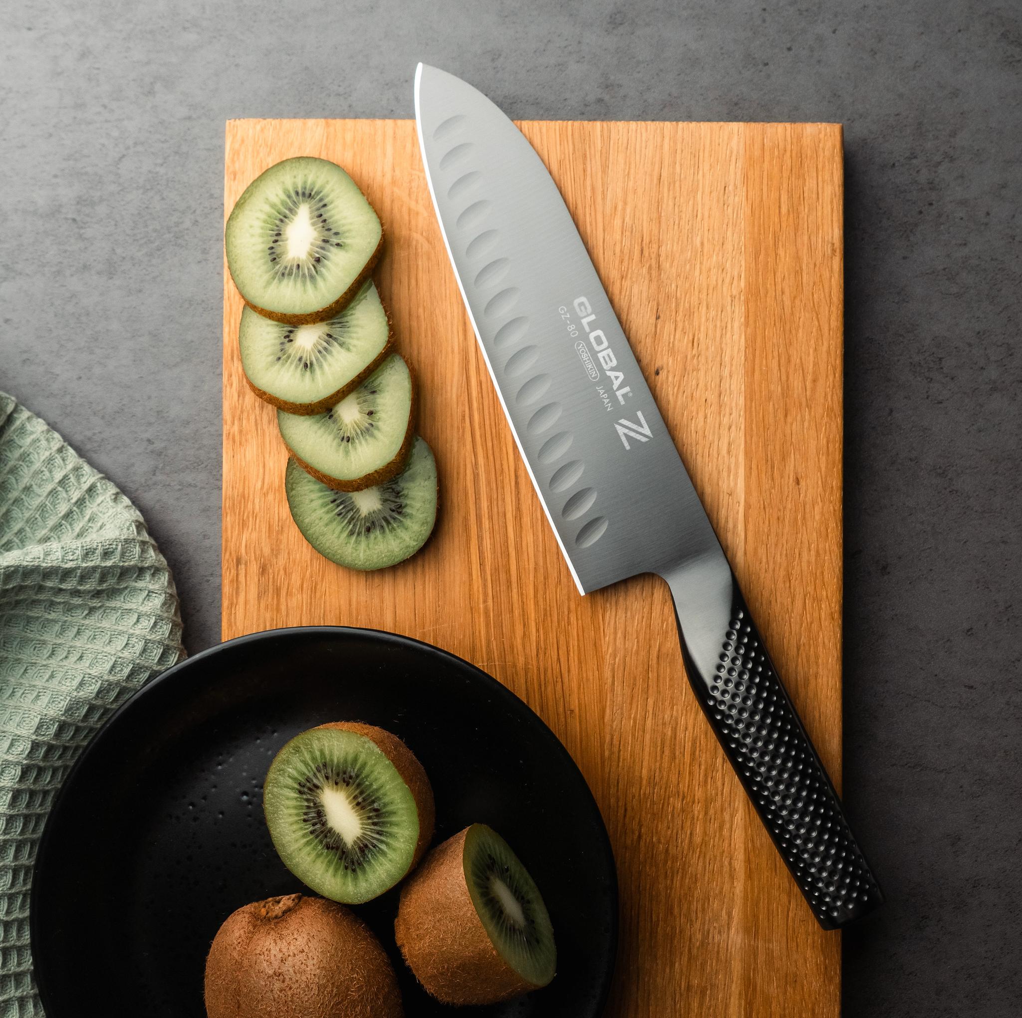 Skivad kiwi och en Global-kökskniv på en skärbräda bredvid en svart tallrik med färska kiwifrukter.