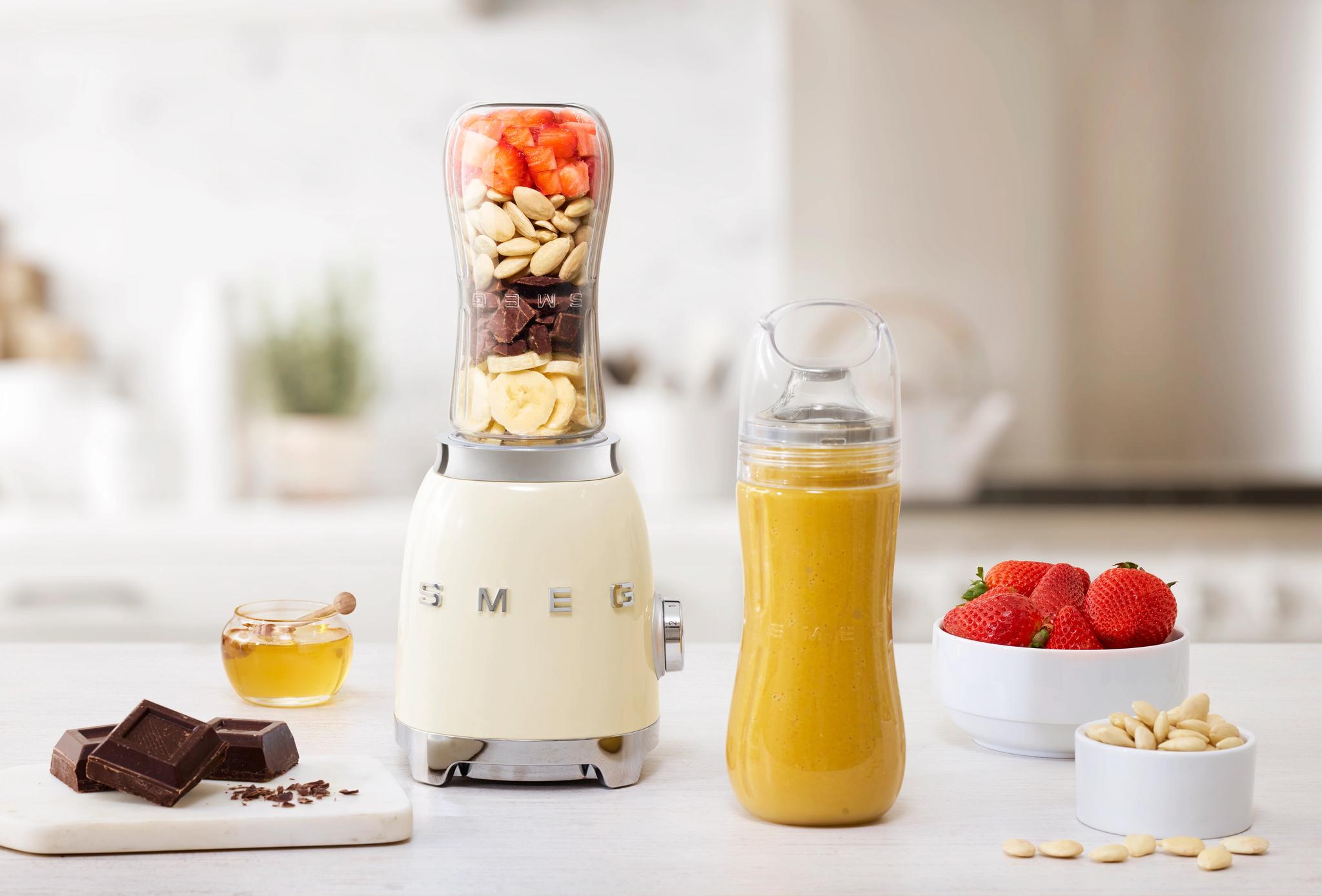 En krämfärgad Smeg-mixer med lager av jordgubbar, mandlar, choklad och banan, bredvid en gul smoothie i en flaska, jordgubbar, mandlar, choklad och honung på en vit köksbänk.