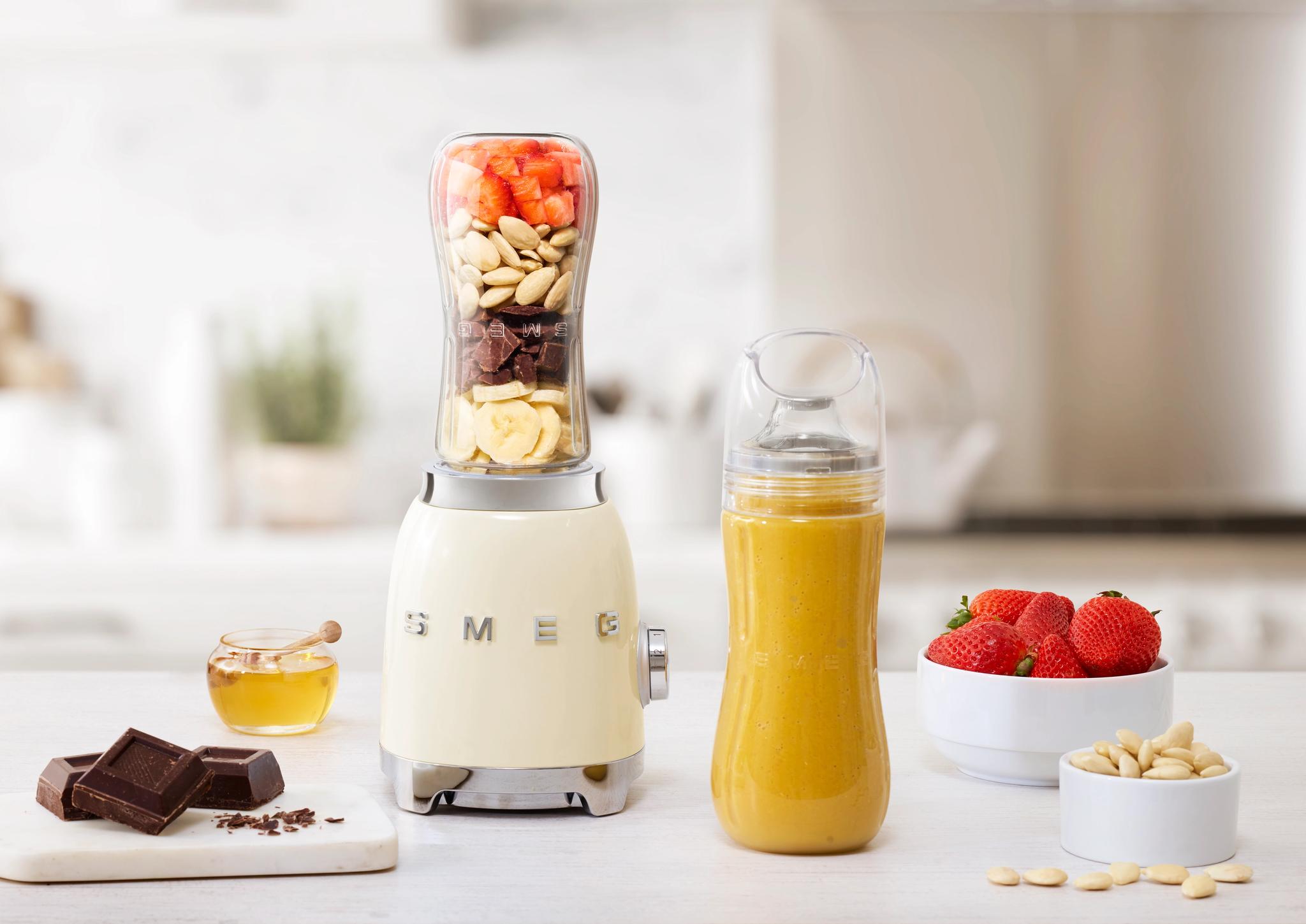 Kermainen Smeg-tehosekoitin, jonka päällä on kerroksia banaania, suklaata, manteleita ja mansikoita, keltaisen smoothiepullon, mansikoiden ja suklaapalleroiden vieressä keittiön tiskillä.