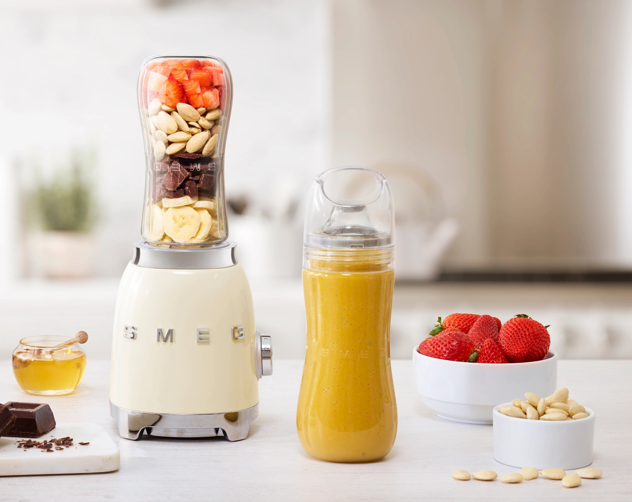 En krämfärgad Smeg-mixer med lager av jordgubbar, mandlar, choklad och banan, bredvid en gul smoothie i en flaska, jordgubbar, mandlar, choklad och honung på en vit köksbänk.