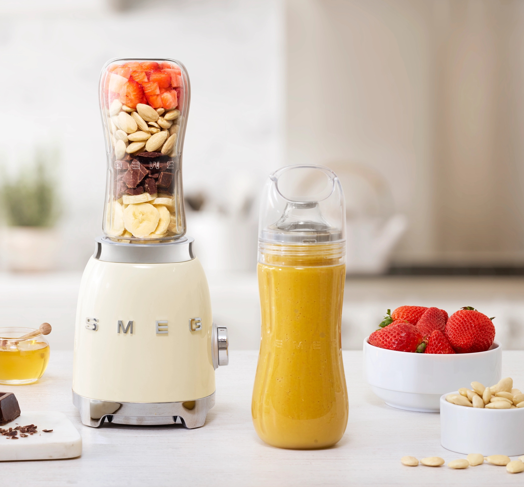 Kermainen Smeg-tehosekoitin, jonka päällä on kerroksia banaania, suklaata, manteleita ja mansikoita, keltaisen smoothiepullon, mansikoiden ja suklaapalleroiden vieressä keittiön tiskillä.