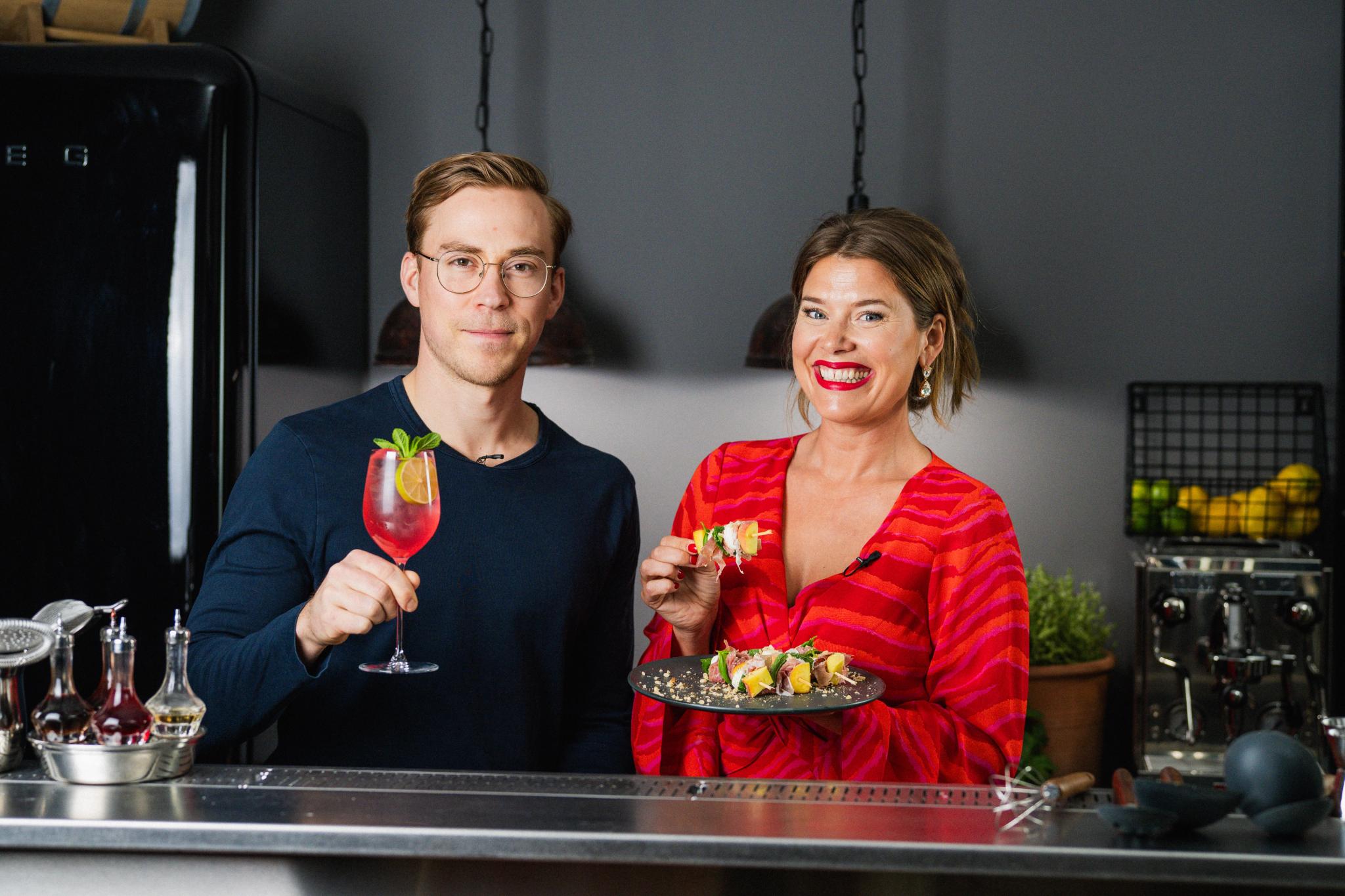 Två leende personer i ett modernt kök håller i en färgglad drink och en tallrik med aptitretare.