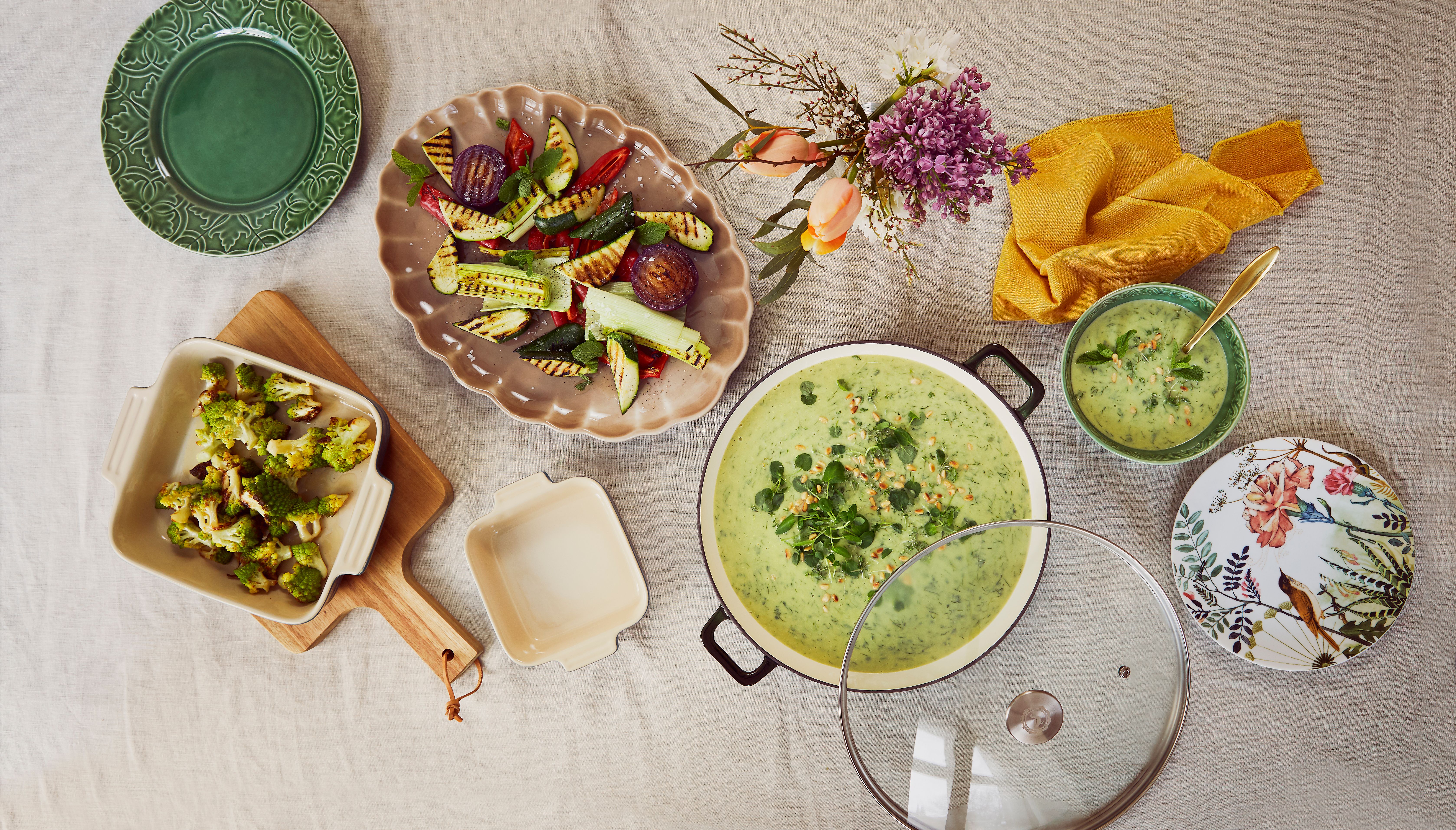 En livfull måltid upplagd på en lätt duk, med en stor gryta med grön soppa, grillade grönsaker, rostad broccoli, diverse tallrikar och en blombukett.