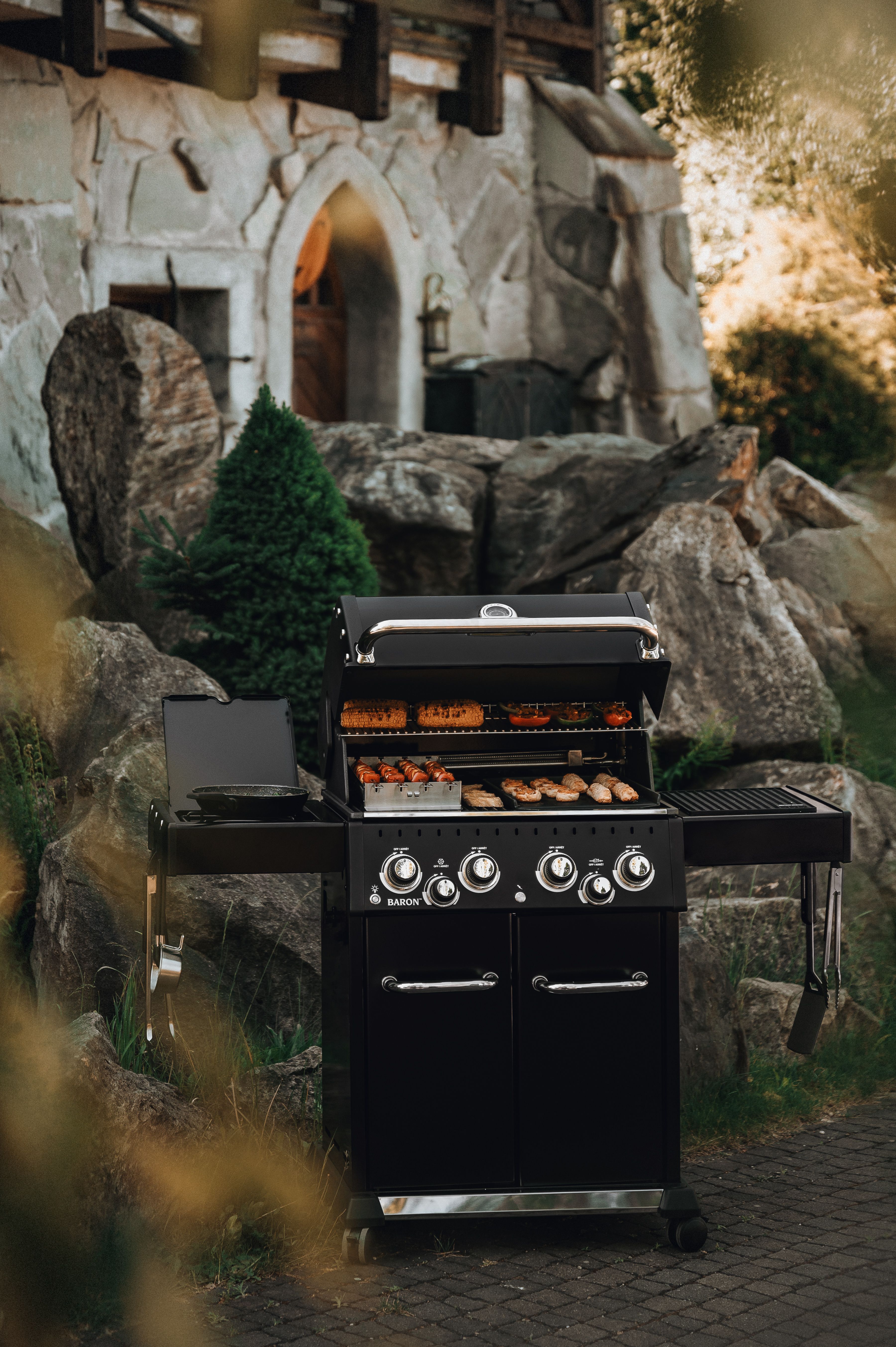 En svart gasolgrill med öppet lock, där mat tillagas, står på en uteplats med en rustik stenbyggnad i bakgrunden