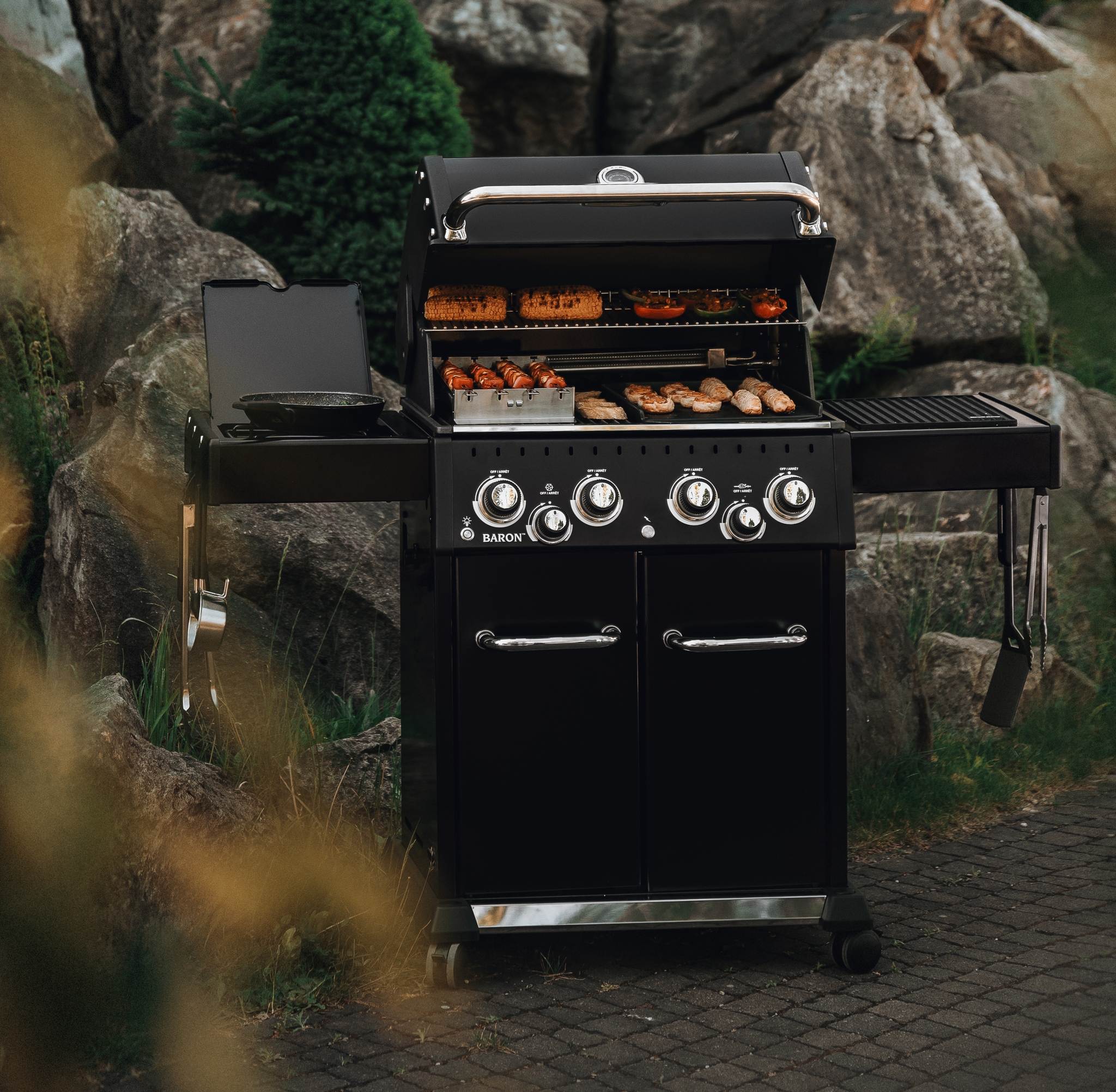 En svart gasolgrill med öppet lock, där mat tillagas, står på en uteplats med en rustik stenbyggnad i bakgrunden