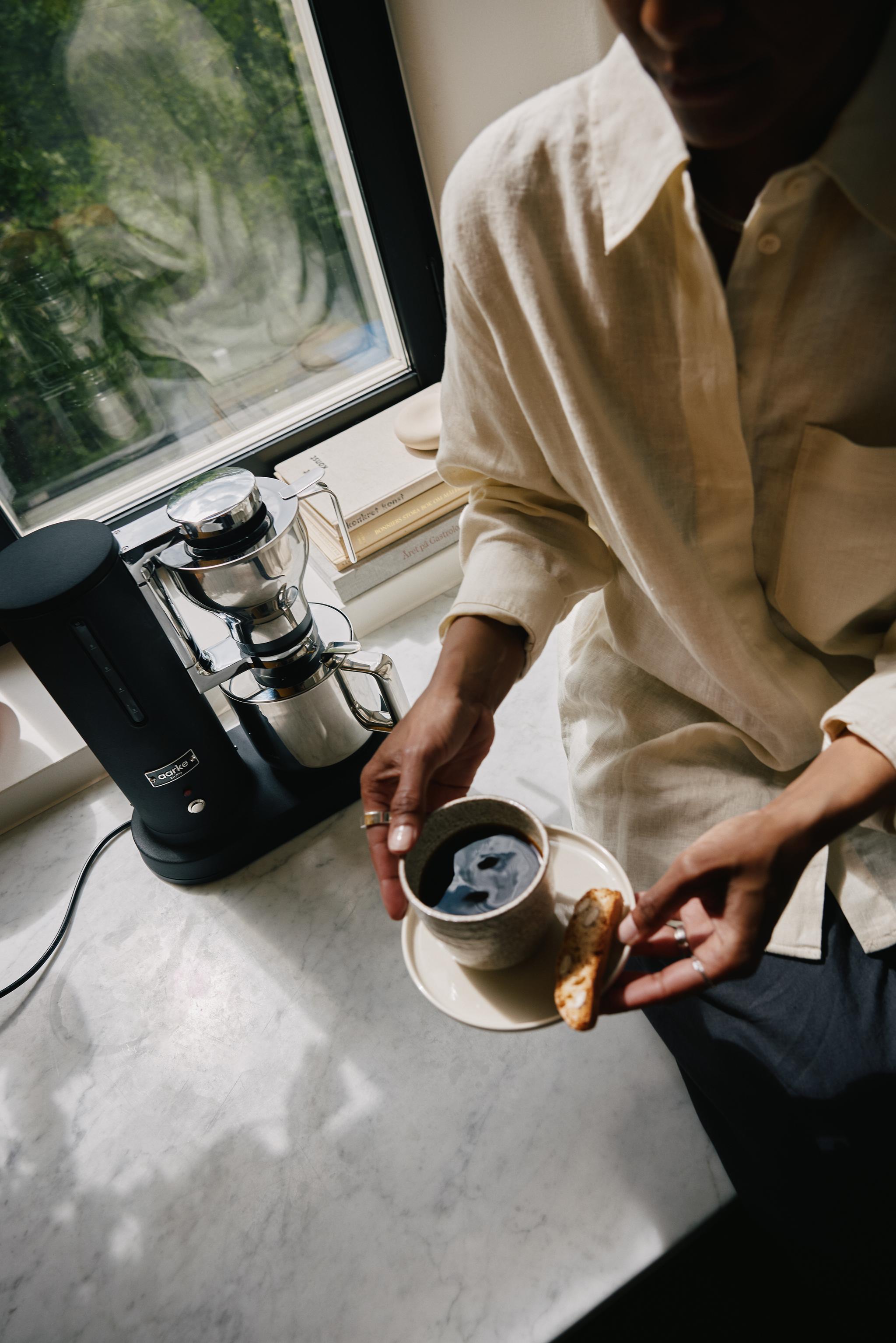 Person som håller en kopp kaffe och en biscotti bredvid en kaffemaskin.