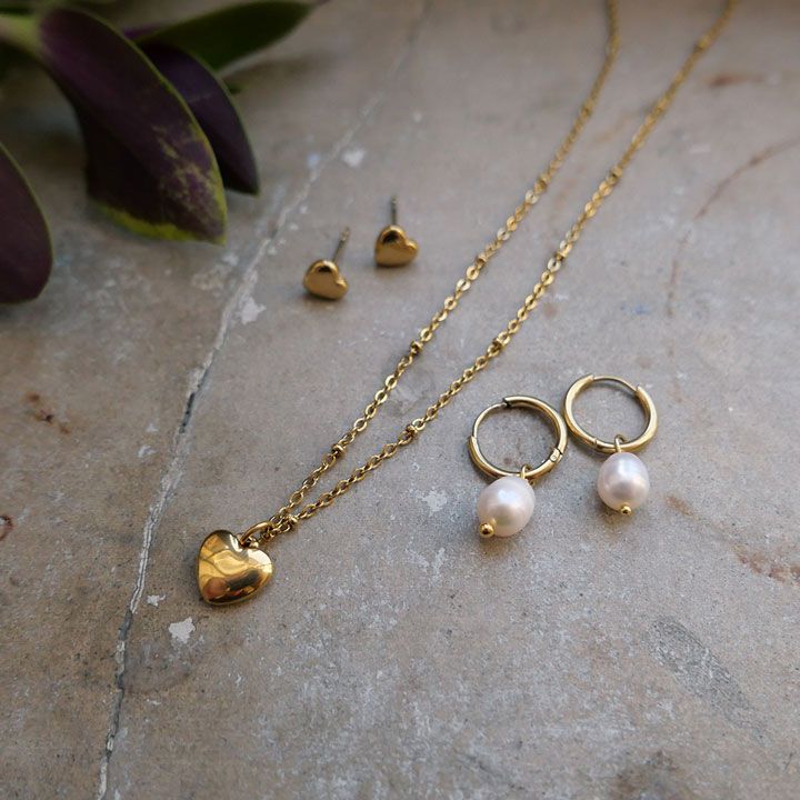 Kultainen kaulakoru sydänriipuksella, sydämenmuotoiset korvakorut ja helmikorvakorut pöydällä