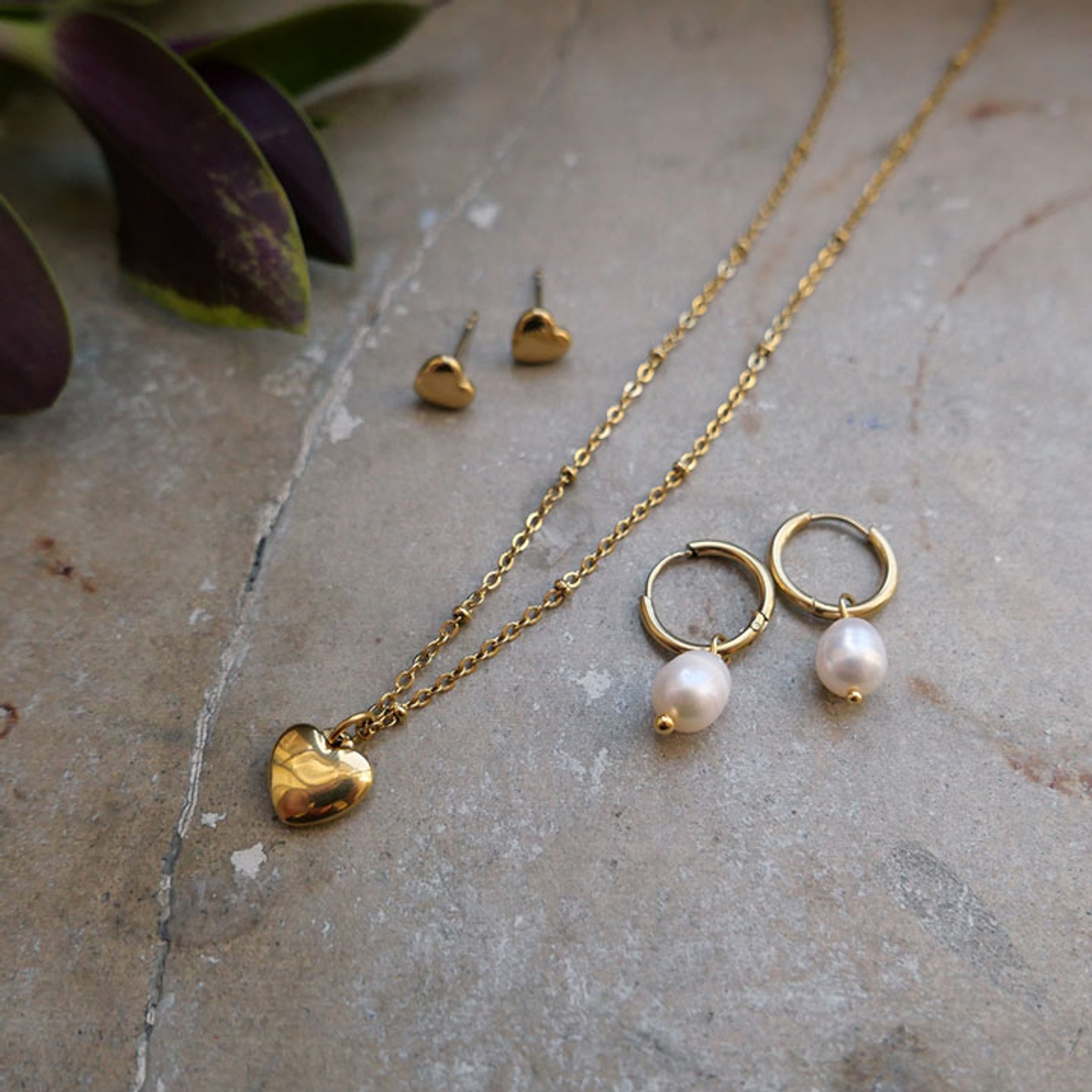 Guldhalsband med hjärthänge, hjärtformade örhängen och pärlörhängen på en stenyta.