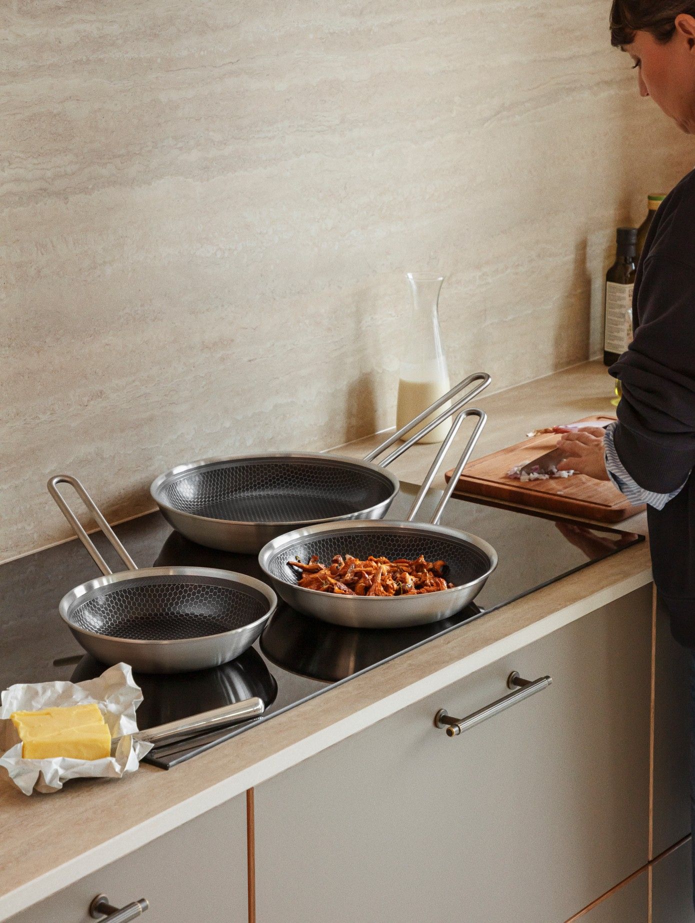 Tre stekpannor i rostfritt stål med bikakemönster på en svart spishäll; en kastrull innehåller kokta svampar, med smör och en stekspade på köksbänken.