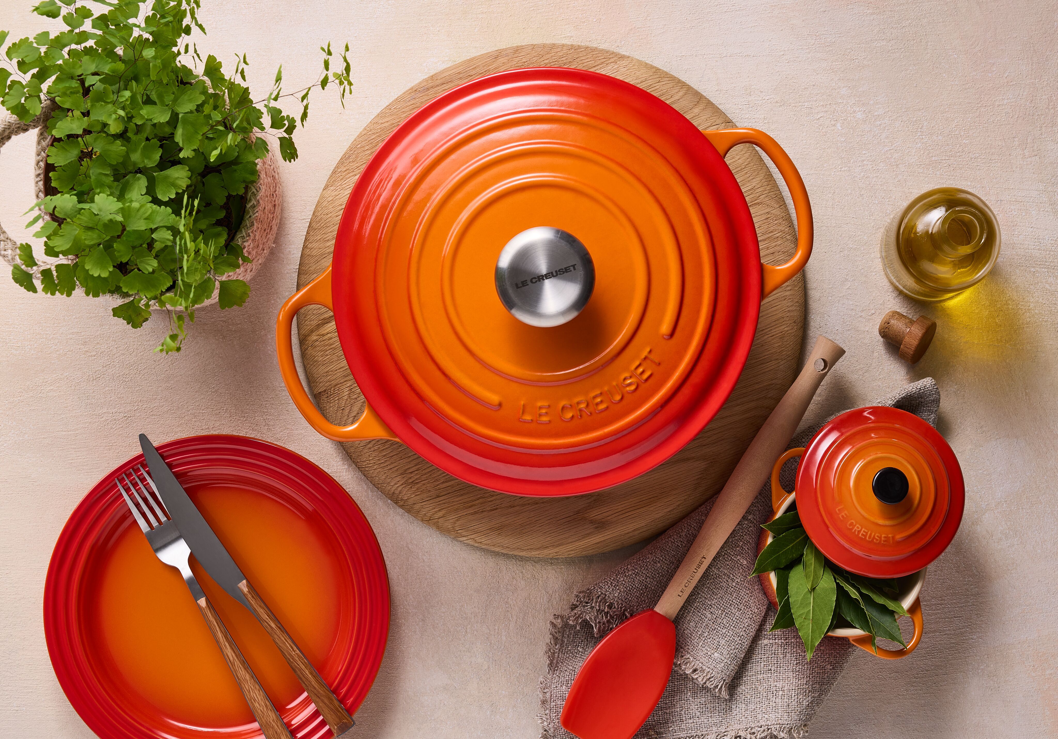 Oranssi Le Creuset -valurautapata ja yhteensopivat astiat kattauksessa keittiön tasolla, vieressä tuoreita yrttejä ja oliiviöljyä.