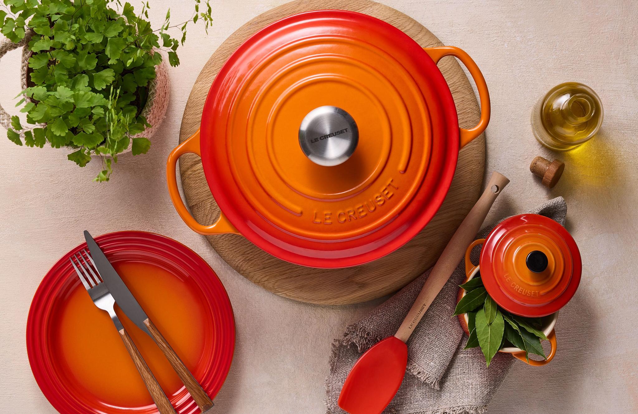 Oranssi Le Creuset -valurautapata ja yhteensopivat astiat kattauksessa keittiön tasolla, vieressä tuoreita yrttejä ja oliiviöljyä.