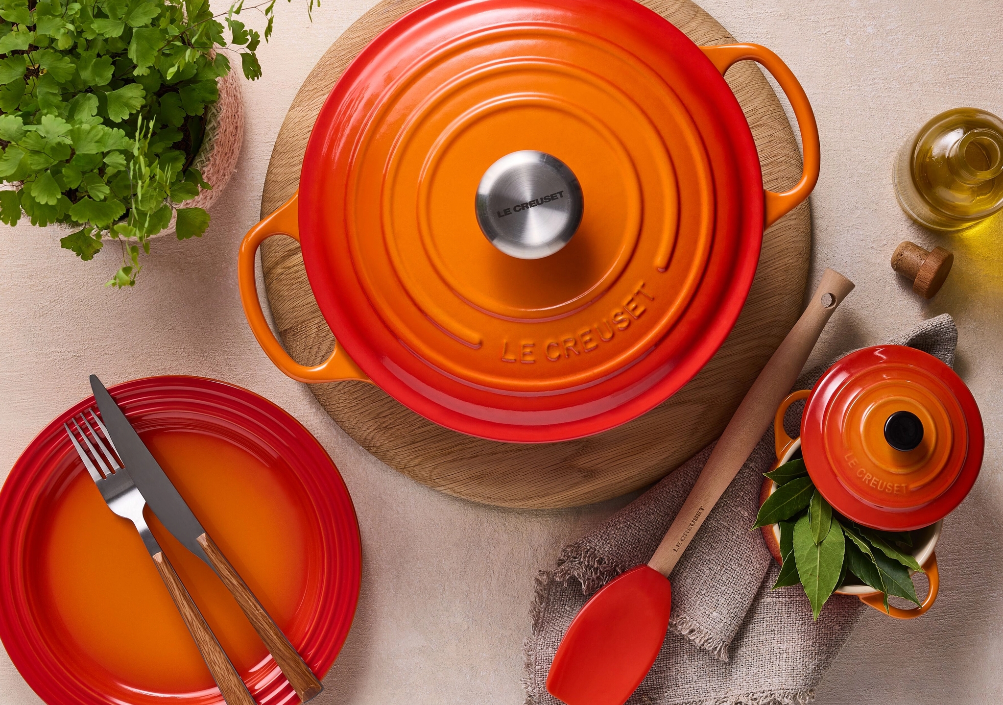 Oranssi Le Creuset -pata, minikattila, lautanen, oliiviöljyä ja kasvi aseteltuna vaalealle taustalle.