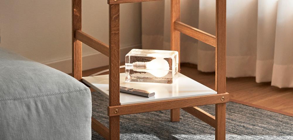 Kaksikerroksinen puinen sivupöytä, jonka päällä on maljakko ja kulho sekä alimmalla hyllyllä ainutlaatuinen kirkas pöytävalaisin.