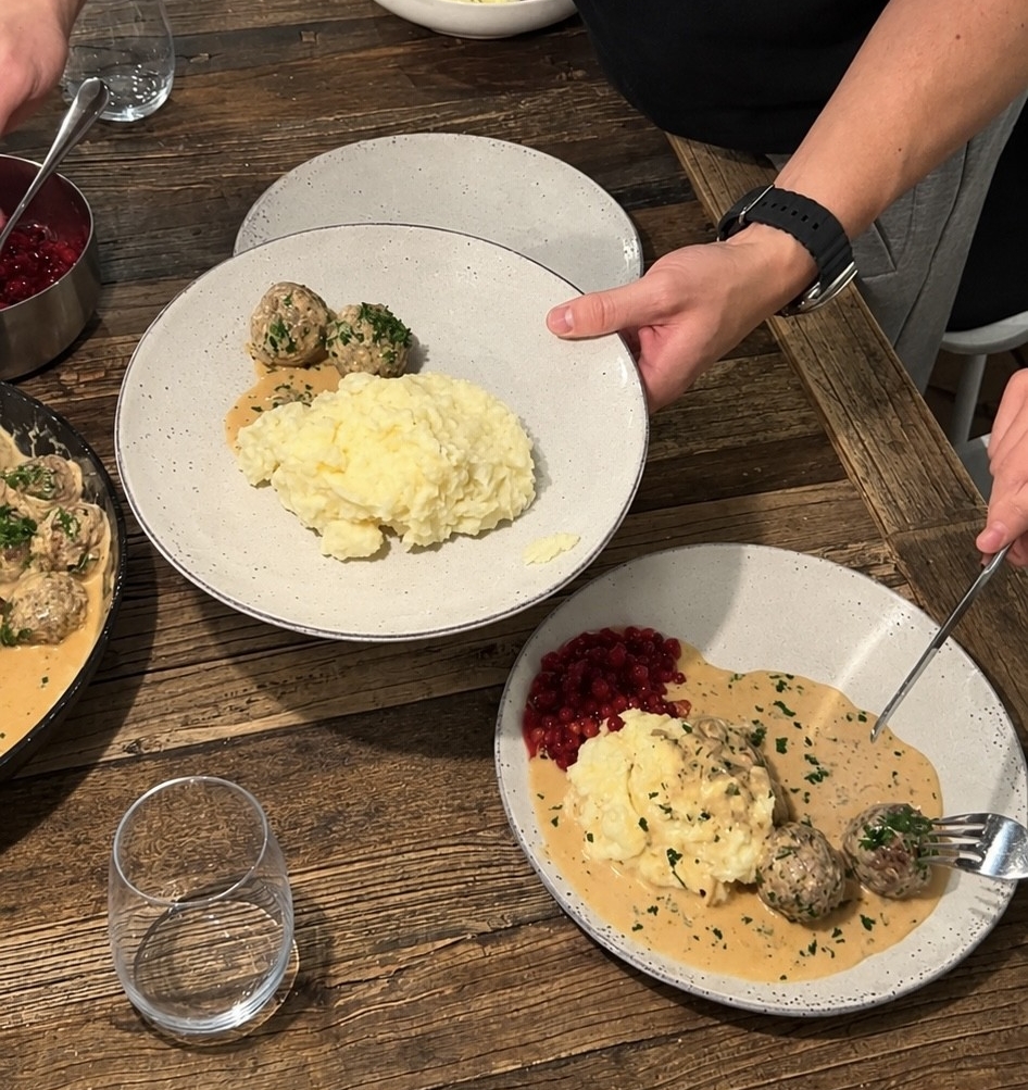 Person serverar svenska köttbullar med potatismos, gräddsås och lingon på ett rustikt träbord.