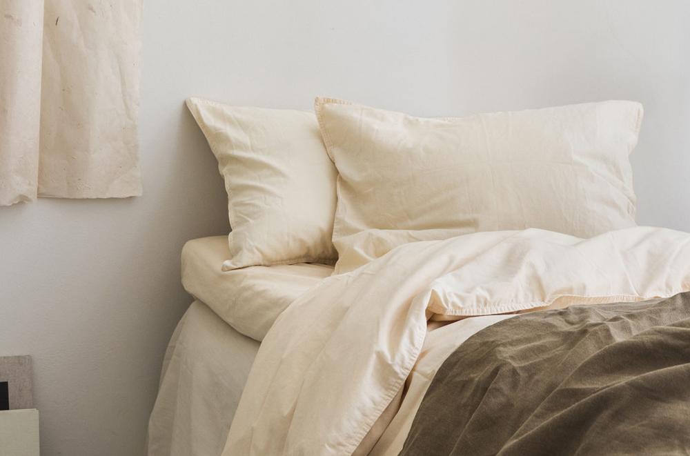 Lyst og minimalistisk soverom med hvitt sengetøy, beige puter og en brun sengepledd.
