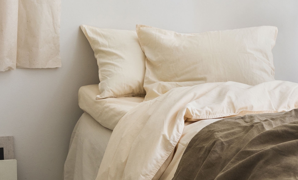 Lyst og minimalistisk soverom med hvitt sengetøy, beige puter og en brun sengepledd.