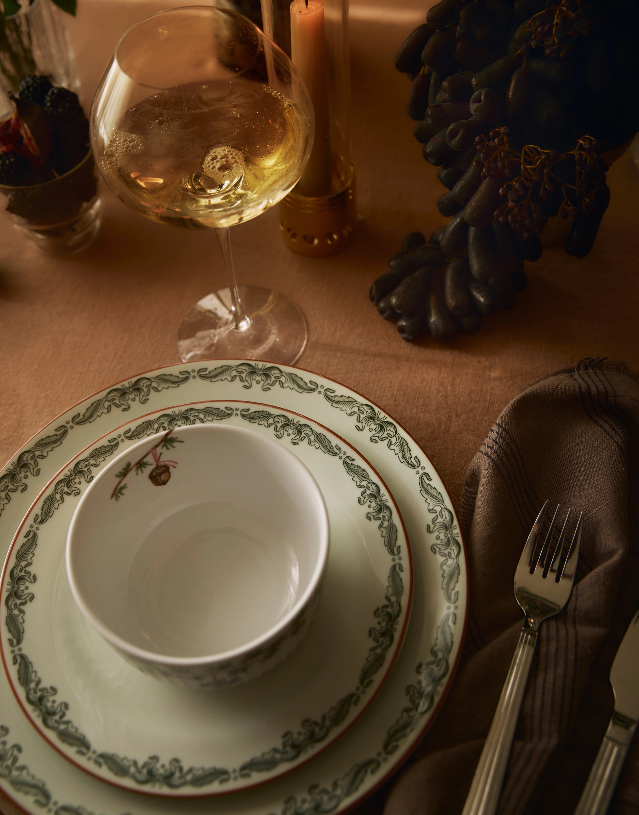 Ruokapöytä, jossa on vihreäkuvioiset lautaset, aterimet, lasi kuohuviiniä, palava kynttilä ja tummia viinirypäleitä.