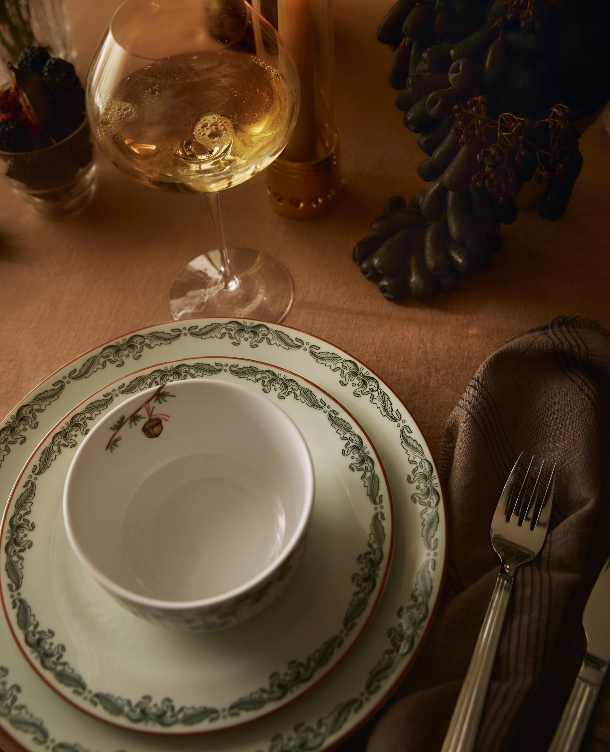 Ruokapöytä, jossa on vihreäkuvioiset lautaset, aterimet, lasi kuohuviiniä, palava kynttilä ja tummia viinirypäleitä.