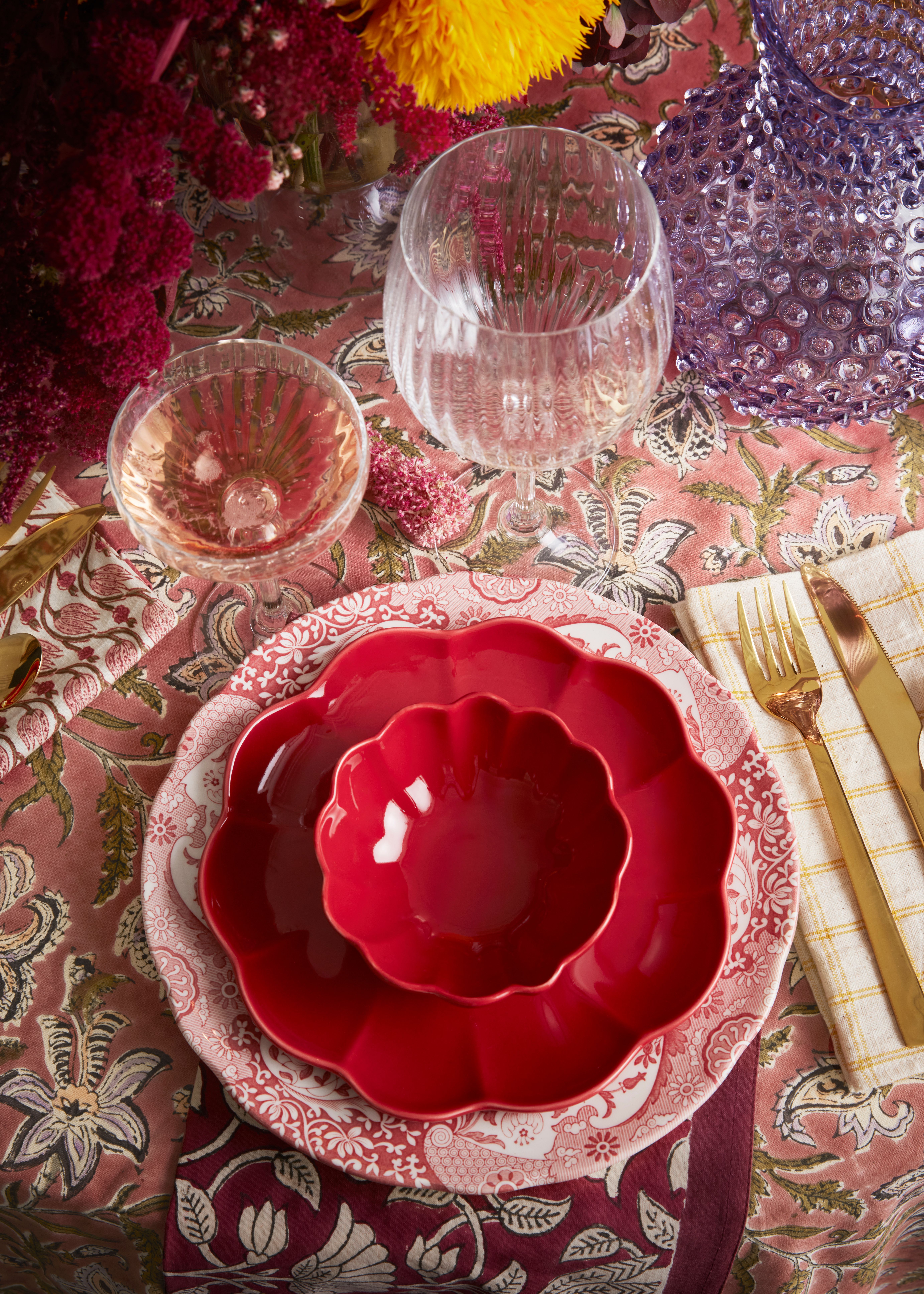 Fargerik borddekking med røde, kamskjellformede tallerkener, vinglass, gullbestikk og fargerike blomster på en mønstret rosa duk.