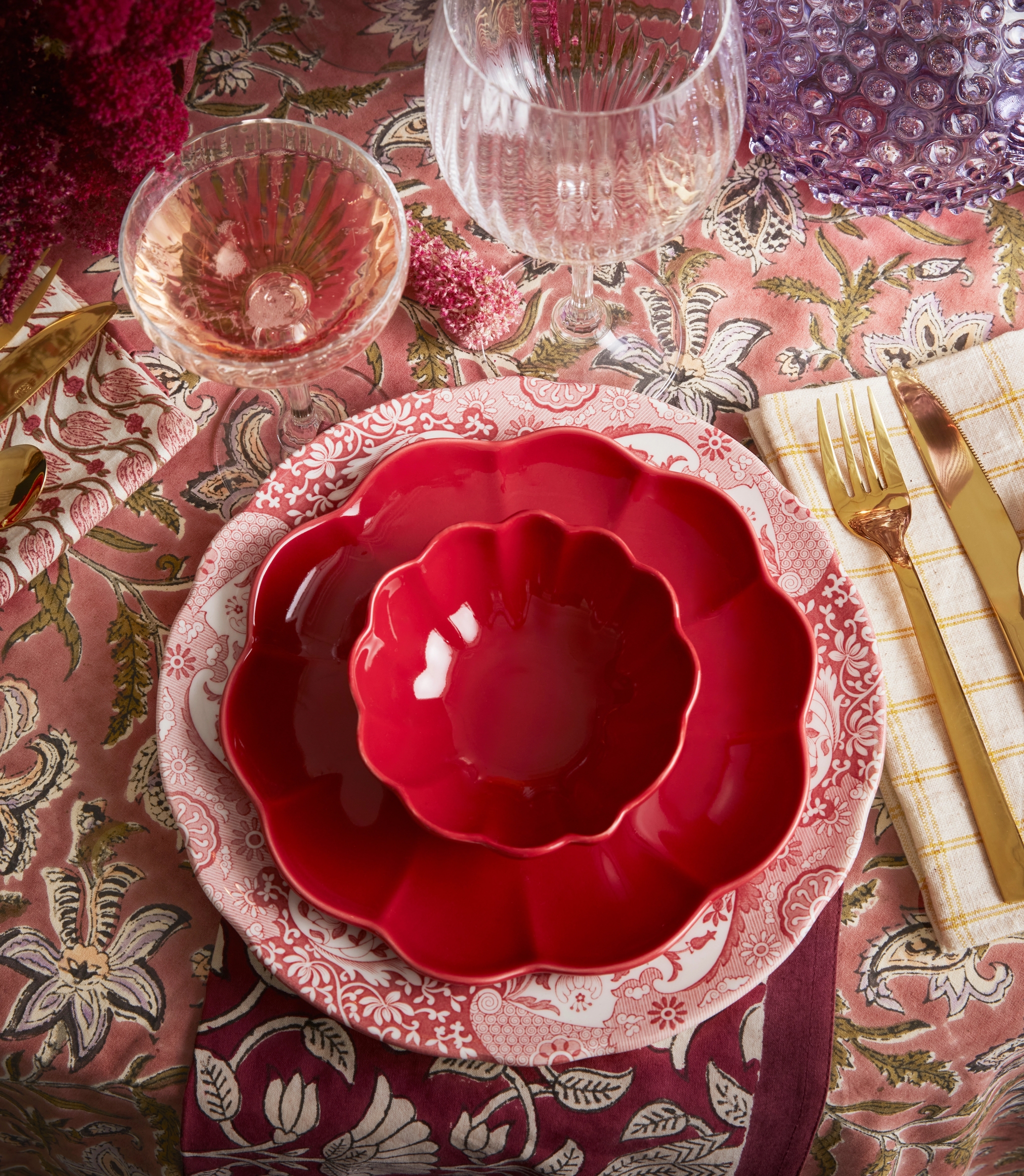 Fargerik borddekking med røde, kamskjellformede tallerkener, vinglass, gullbestikk og fargerike blomster på en mønstret rosa duk.