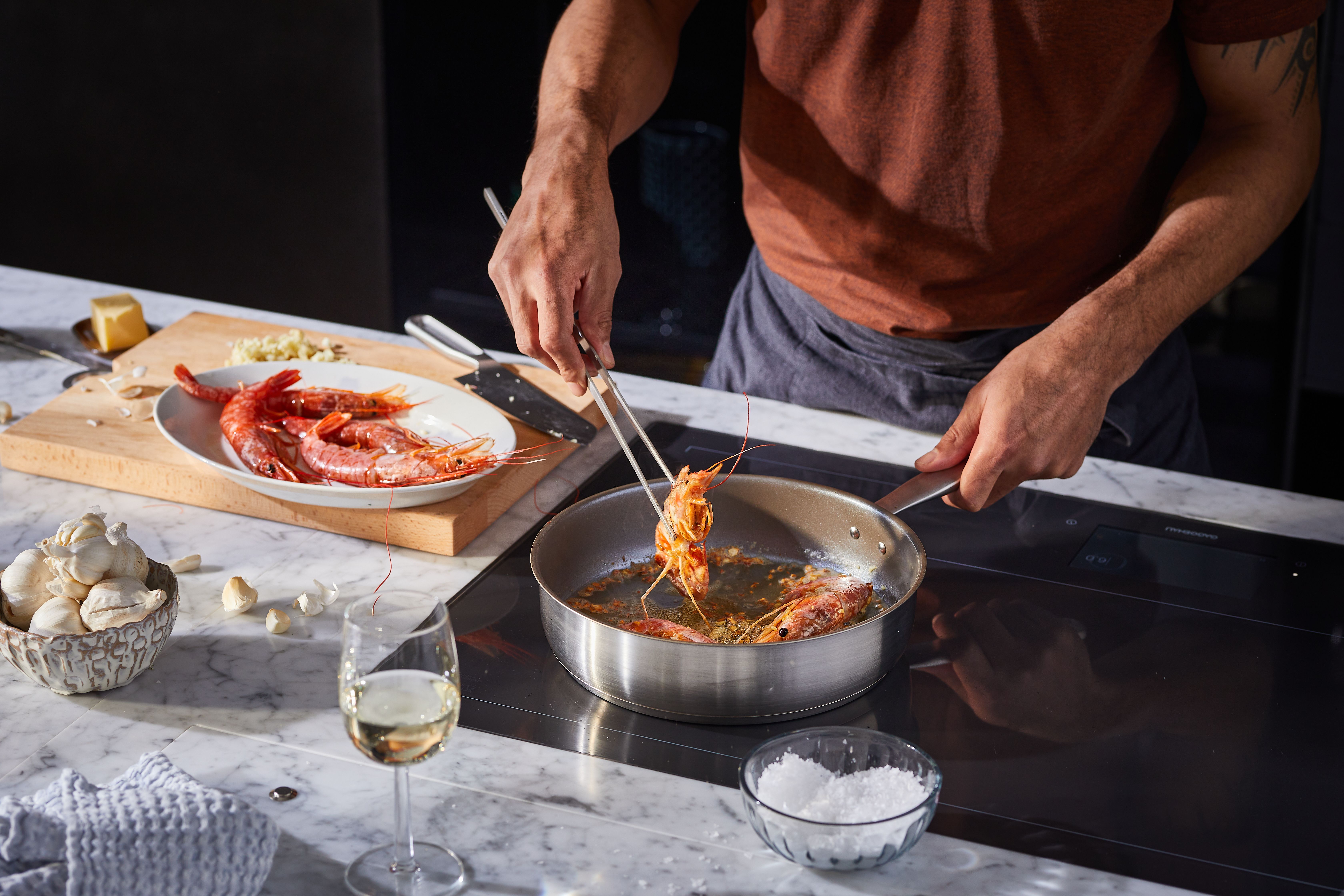 Henkilö kypsentää katkarapuja pannulla pihteillä. Marmorisella tiskillä on raakoja katkarapuja, valkosipulia ja lasillinen viiniä.