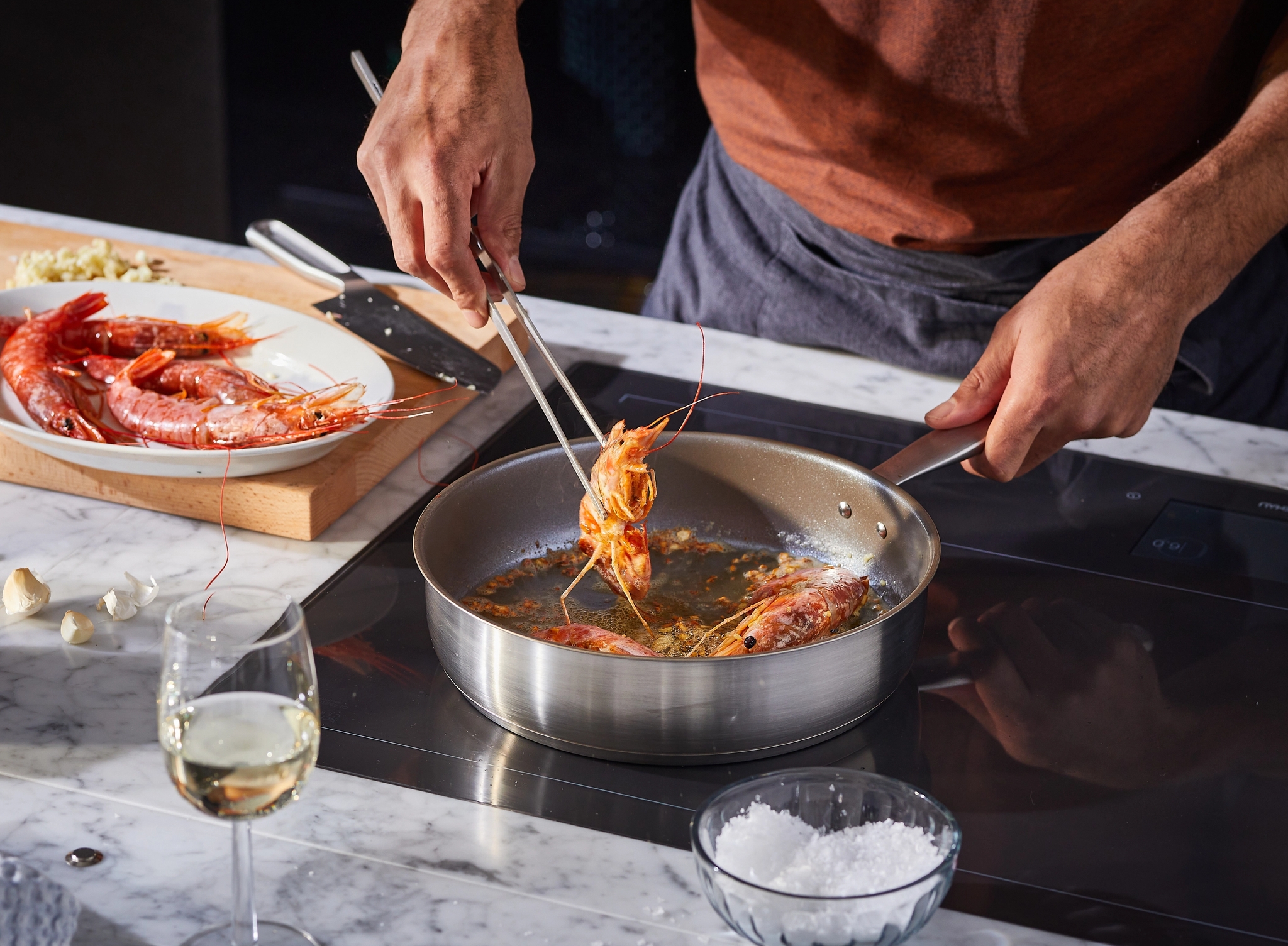 Henkilö kypsentää katkarapuja pannulla pihteillä. Marmorisella tiskillä on raakoja katkarapuja, valkosipulia ja lasillinen viiniä.