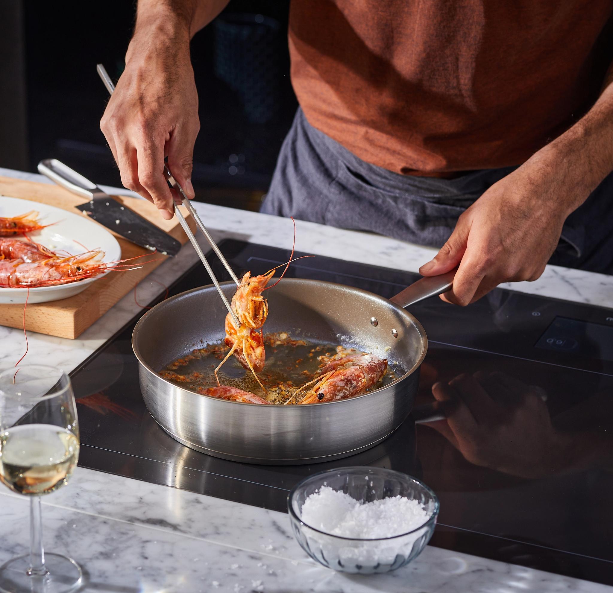 Henkilö kypsentää katkarapuja pannulla pihteillä. Marmorisella tiskillä on raakoja katkarapuja, valkosipulia ja lasillinen viiniä.