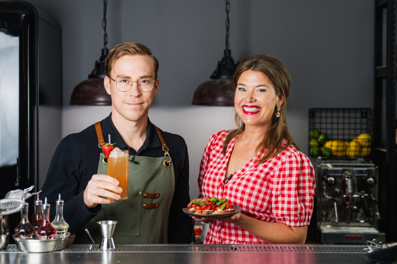 Leende bartender och kvinna i rutig klänning serverar färgglad cocktail och tallrik med mat i en modern bar.