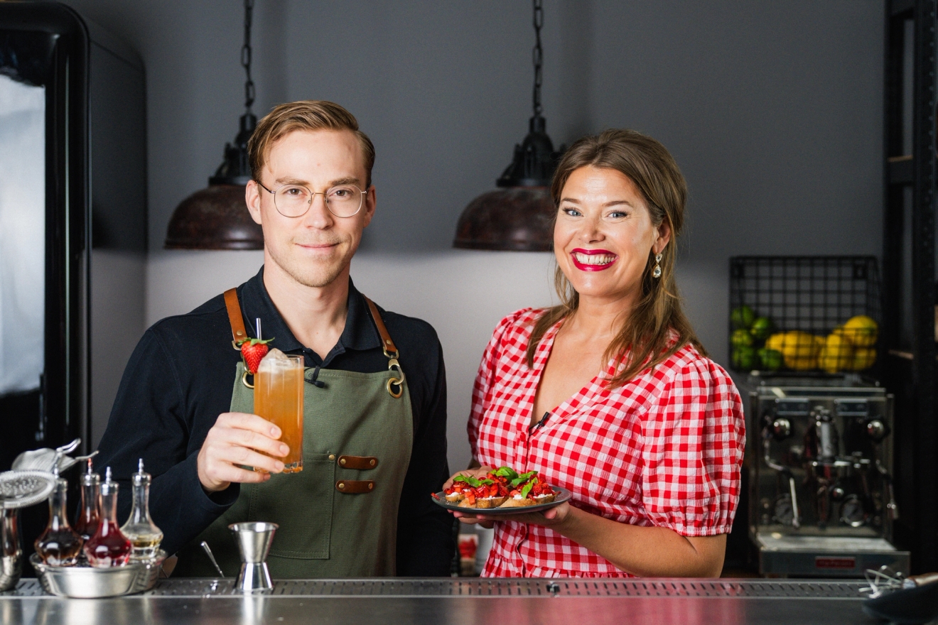 En bartender och en kvinna i rutig klänning står i en bar och visar upp en färgglad cocktail och en tallrik med mat.