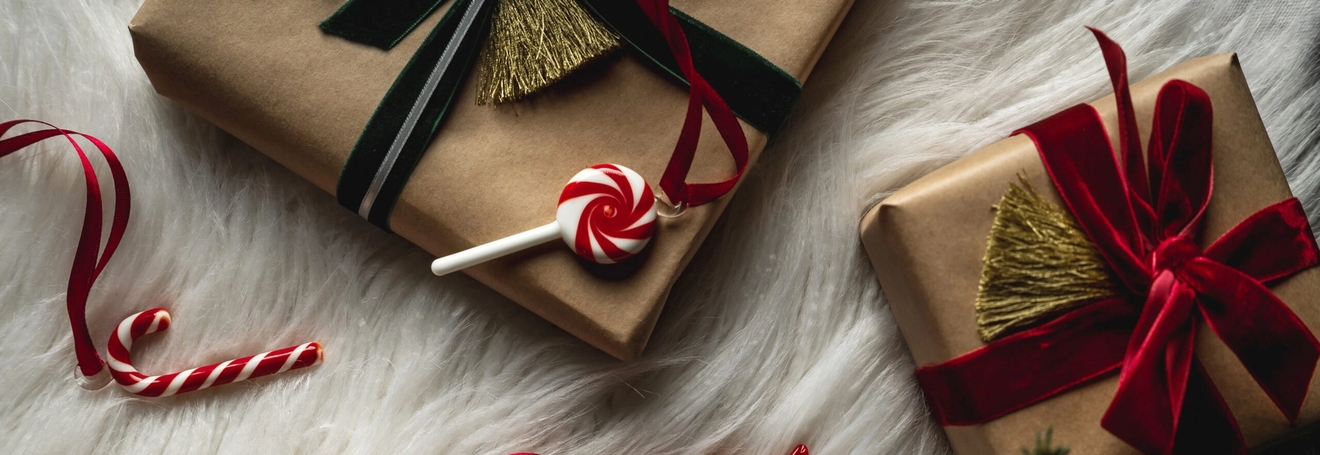 Kolme käärittyä joululahjaa, karkkeja ja männynoksia valkoisella karvaisella taustalla.