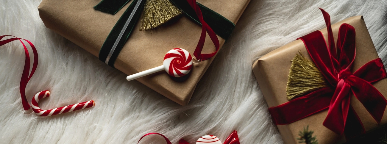 Kolme käärittyä joululahjaa, karkkeja ja männynoksia valkoisella karvaisella taustalla.