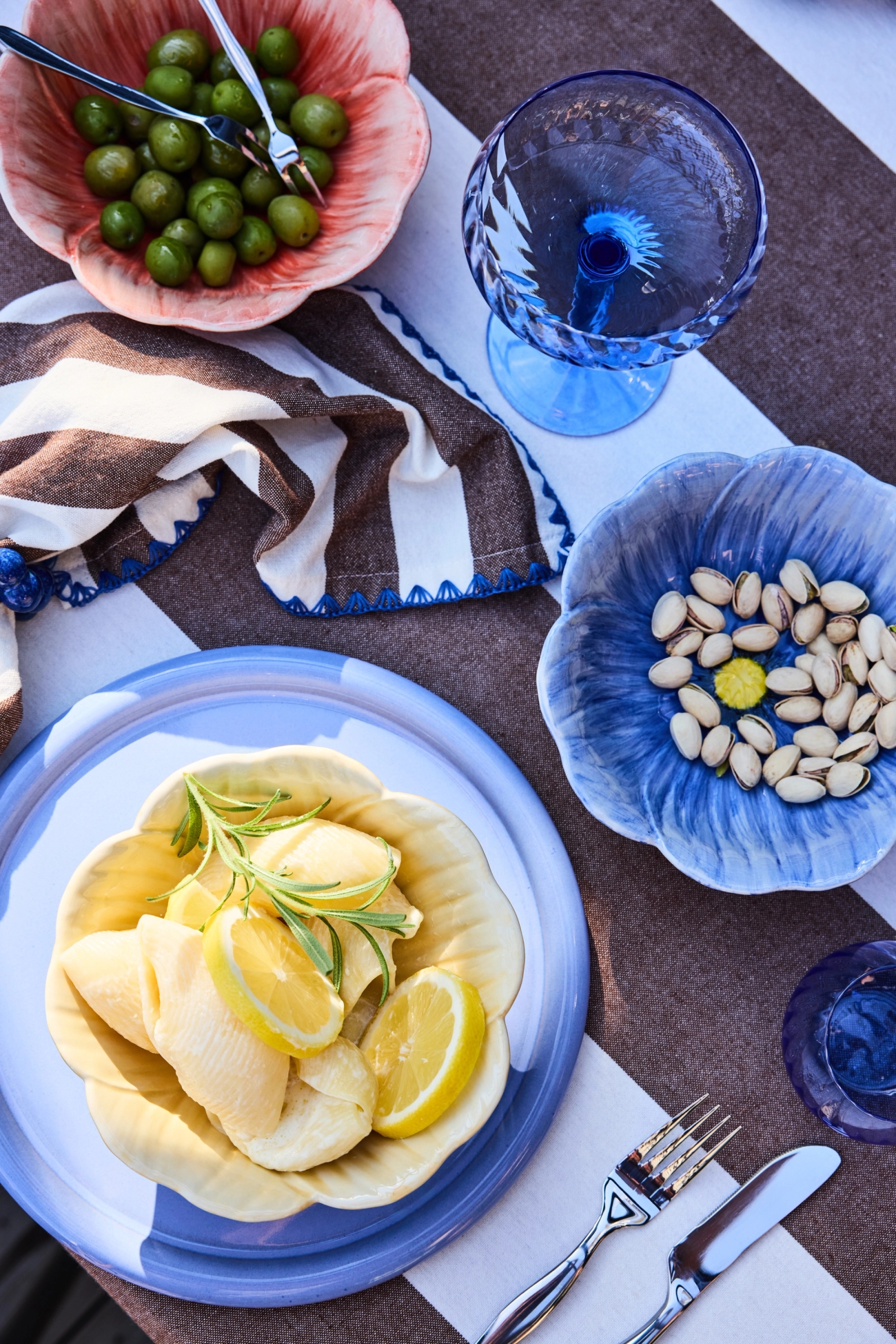 Overhead view of a colorful table setting with flower-shaped bowls of olives, pistachios, and pasta, alongside a blue glass and striped napkin.