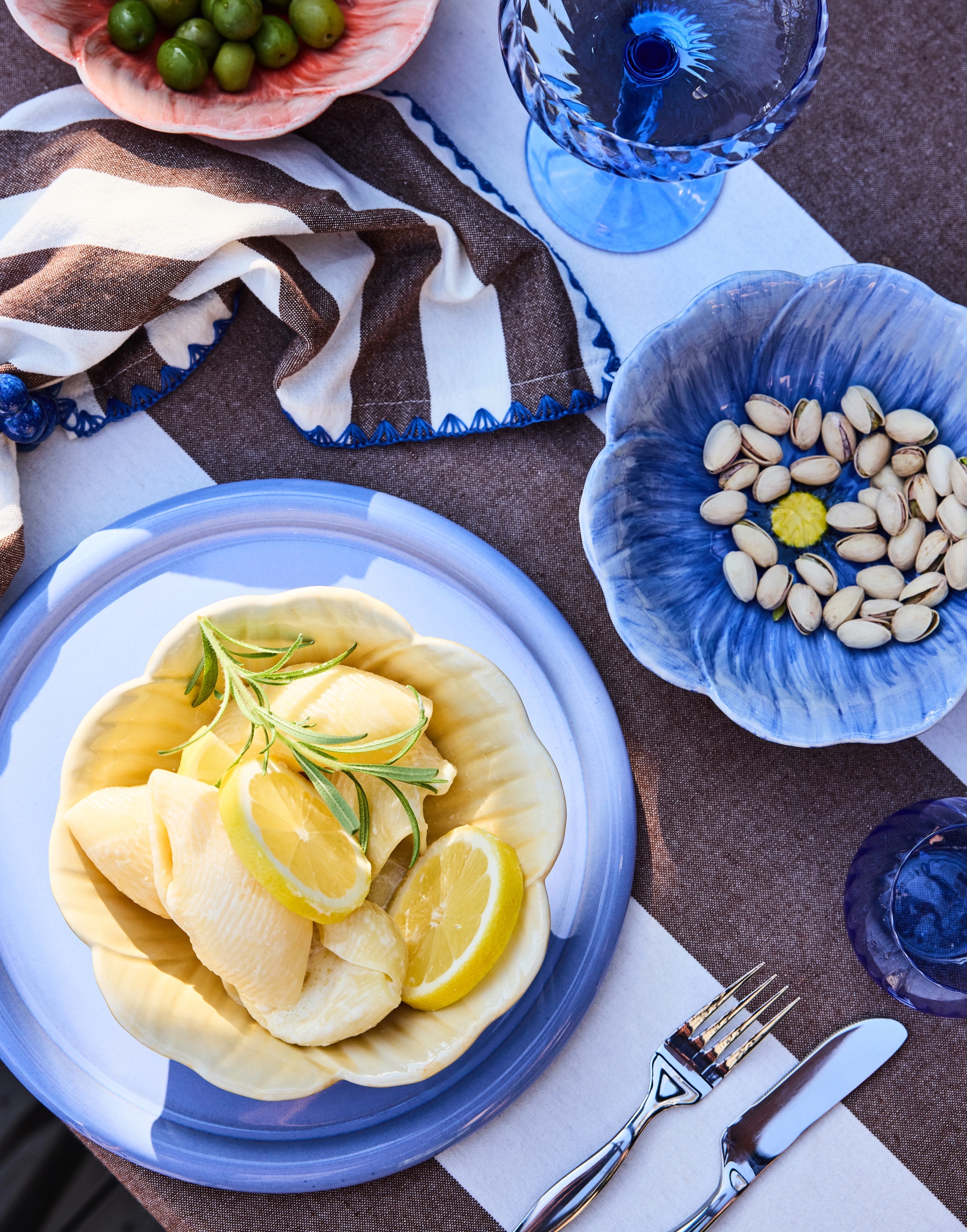 An overhead view of a table set with a brown and white striped cloth, featuring bowls of olives, pistachios, and hearts of palm with lemon and rosemary, along with blue plates and glassware.
