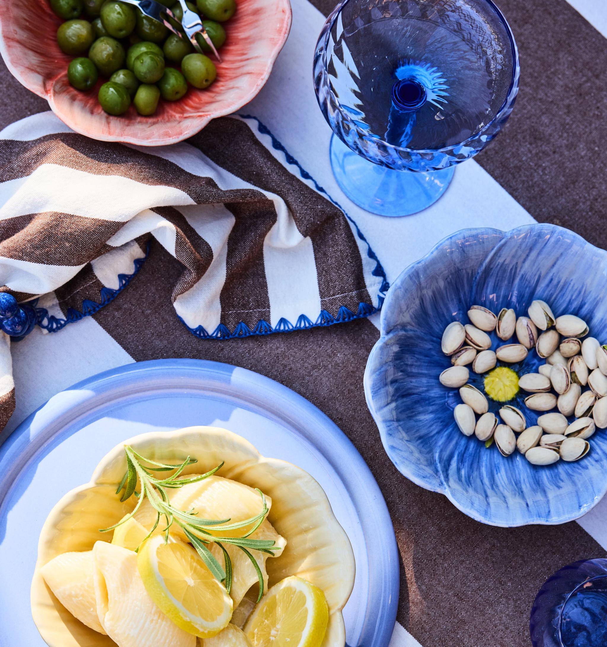 An overhead view of a table set with a striped cloth, featuring floral bowls of green olives, pistachios, and pasta with lemon, alongside a blue glass.