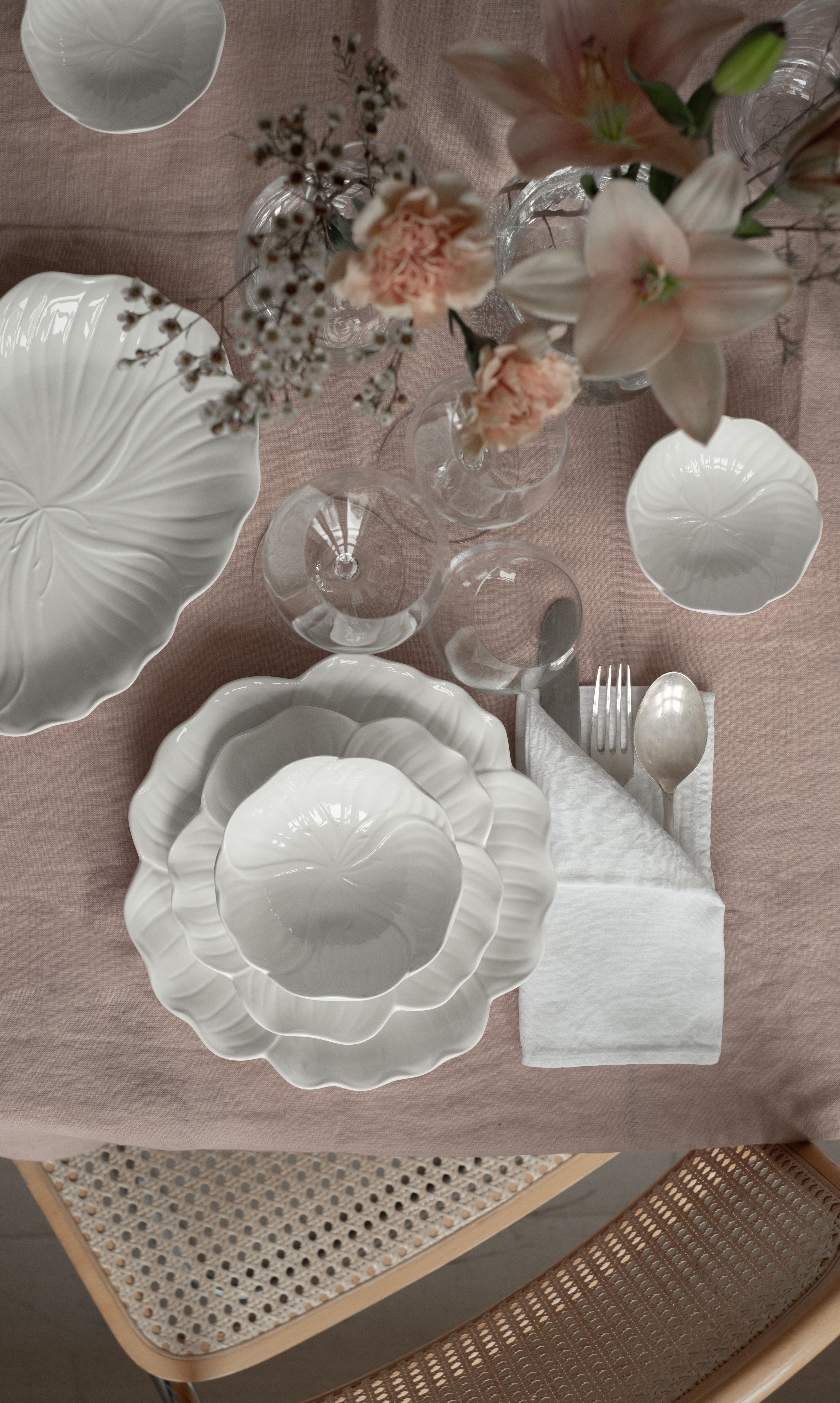 Et ovenfrabilde av et spisebord dekket med hvite kronbladformede tallerkener, klart glass, sølvtøy og rosa blomster på en rødlig duk.