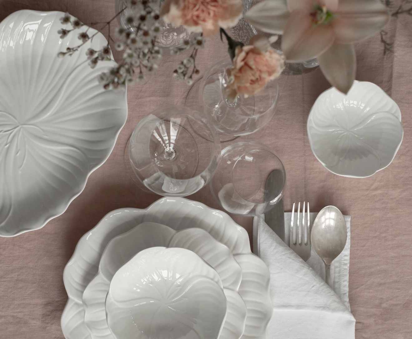 Bild ovanifrån av ett bord dukat med vitt blomformat porslin, genomskinligt glas, bestick och rosa blommor på en ljusrosa duk.