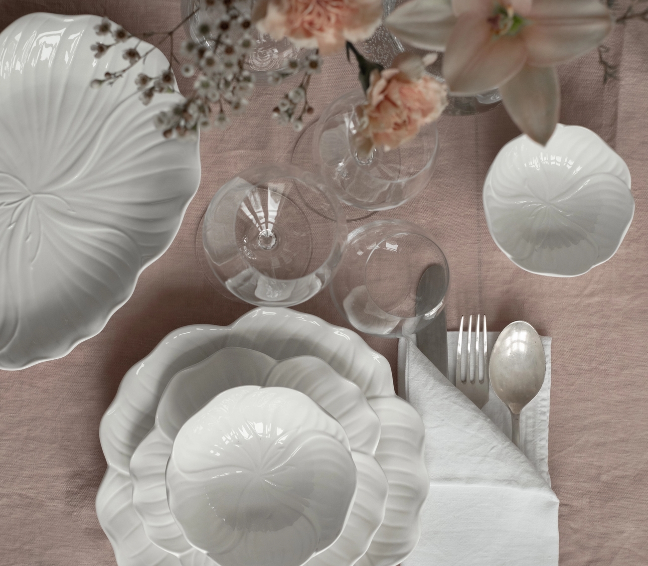 Ovenfra-bilde av et bord dekket med hvitt blomsterformet servise, klart glass, sølvtøy og rosa blomster på en lyserosa duk.