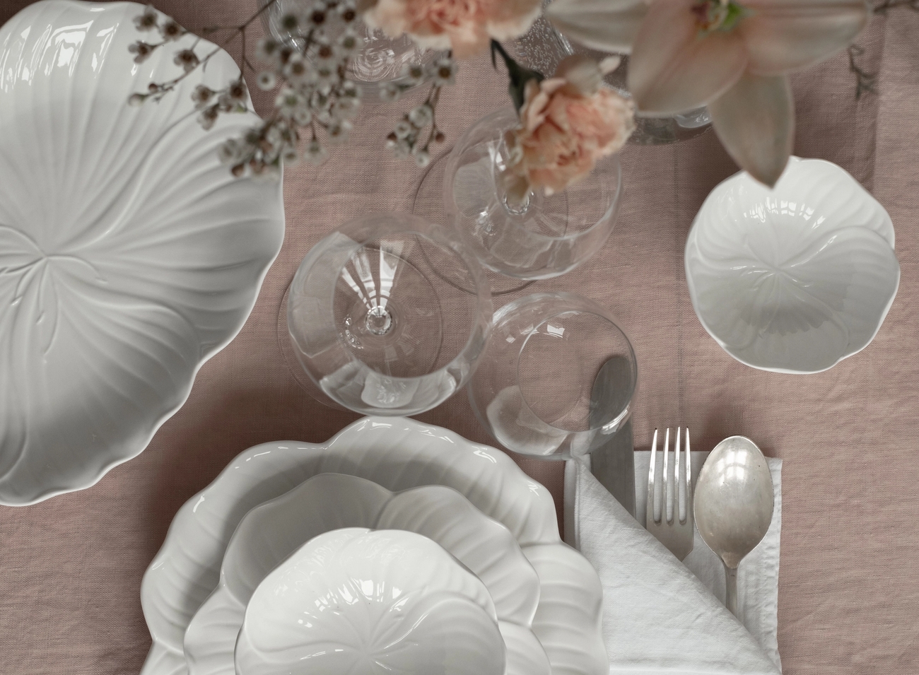 Bild ovanifrån av ett bord dukat med vitt blomformat porslin, genomskinligt glas, bestick och rosa blommor på en ljusrosa duk.
