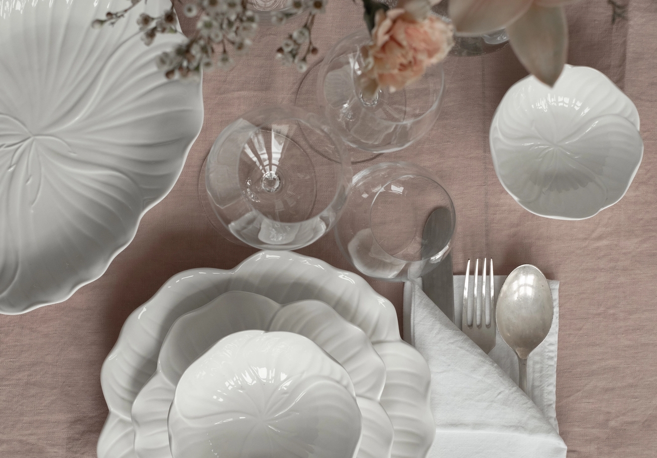 Ovenfra-bilde av et bord dekket med hvitt blomsterformet servise, klart glass, sølvtøy og rosa blomster på en lyserosa duk.