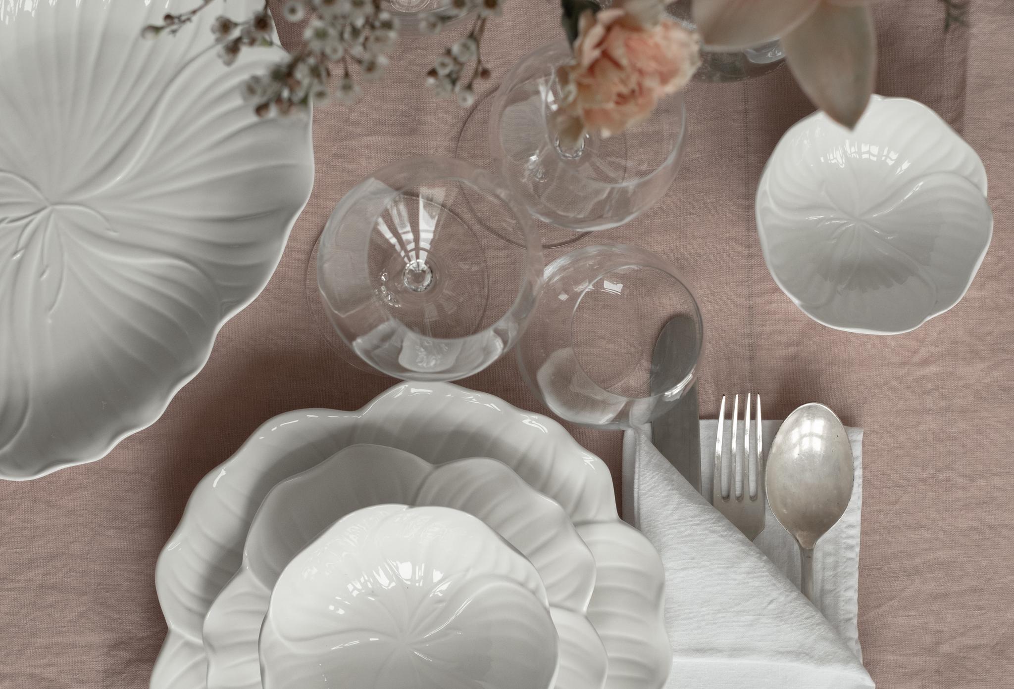 Ovanifrånbild av ett matbord dukat med vitt porslin med bladmotiv, klara glas och bestick. Rosa blommor ligger i en vas på en duk i dammrosa, med en rottingstol längst ner.