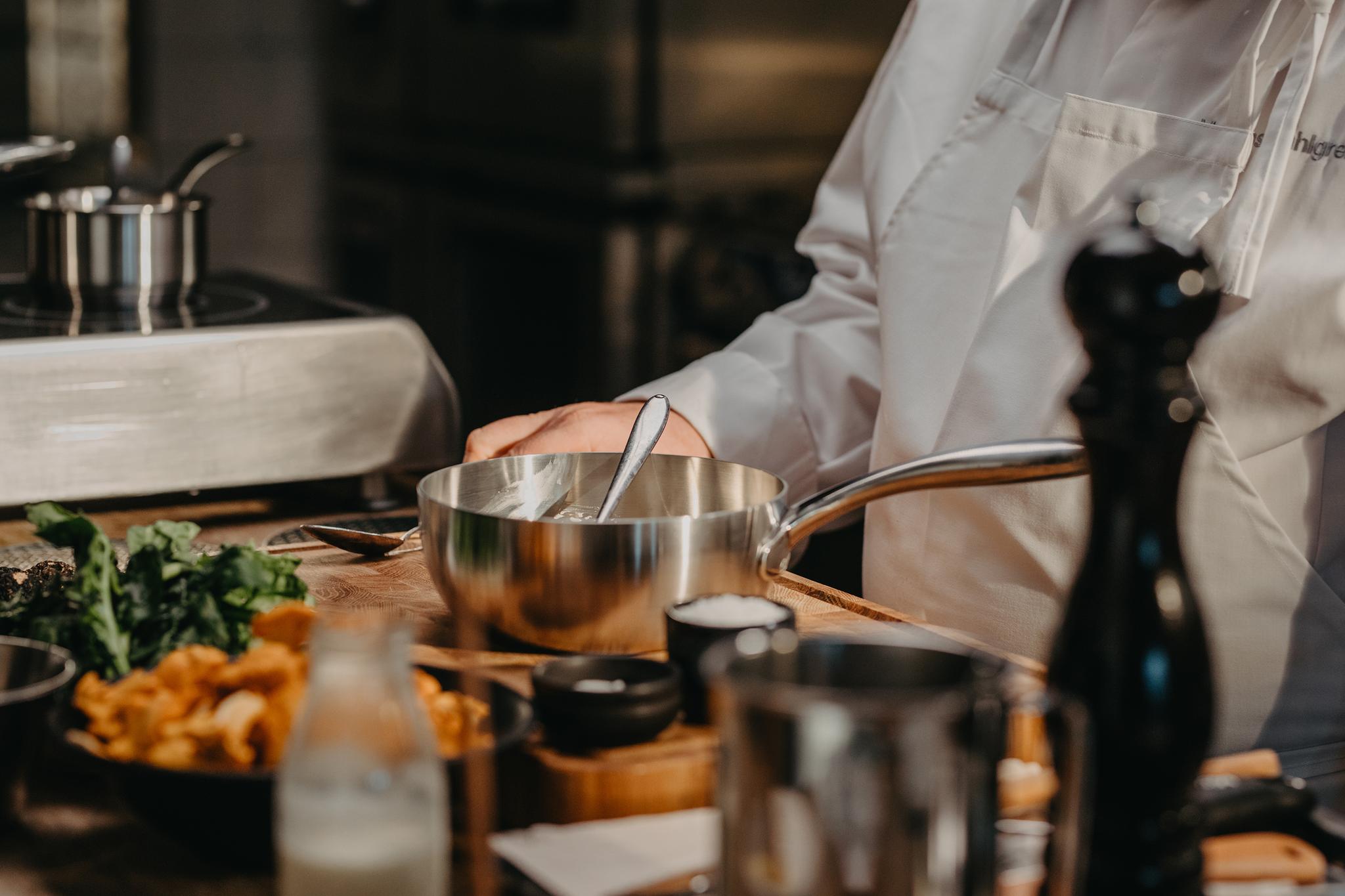 En kock i vit rock rör om ingredienser i en metallskål, omgiven av färska svampar, örter och andra köksredskap på en träbänk.