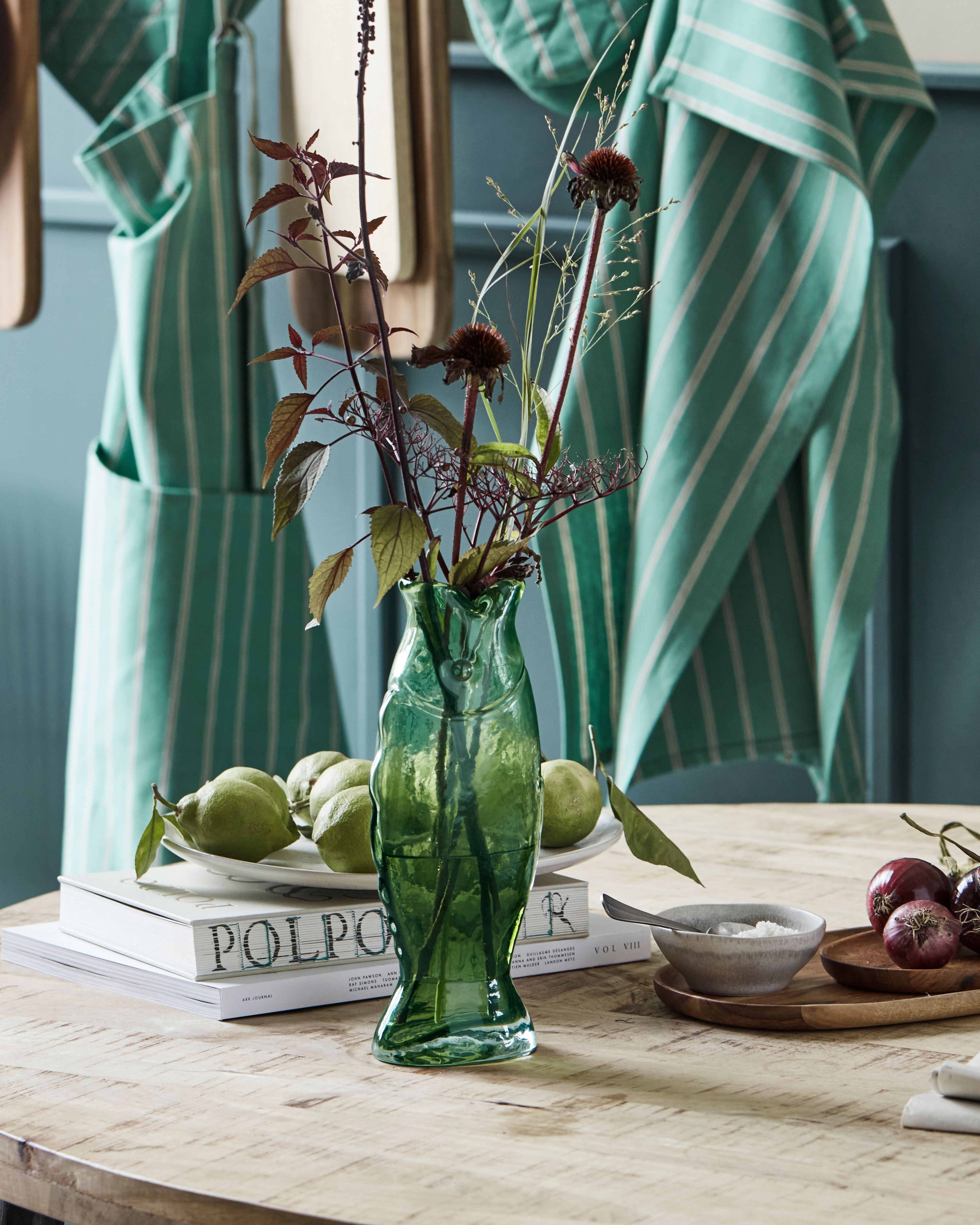 A green fish-shaped vase with a dark floral arrangement sits on a wooden table next to green fruit, red onions, and books, with striped aprons in the background.