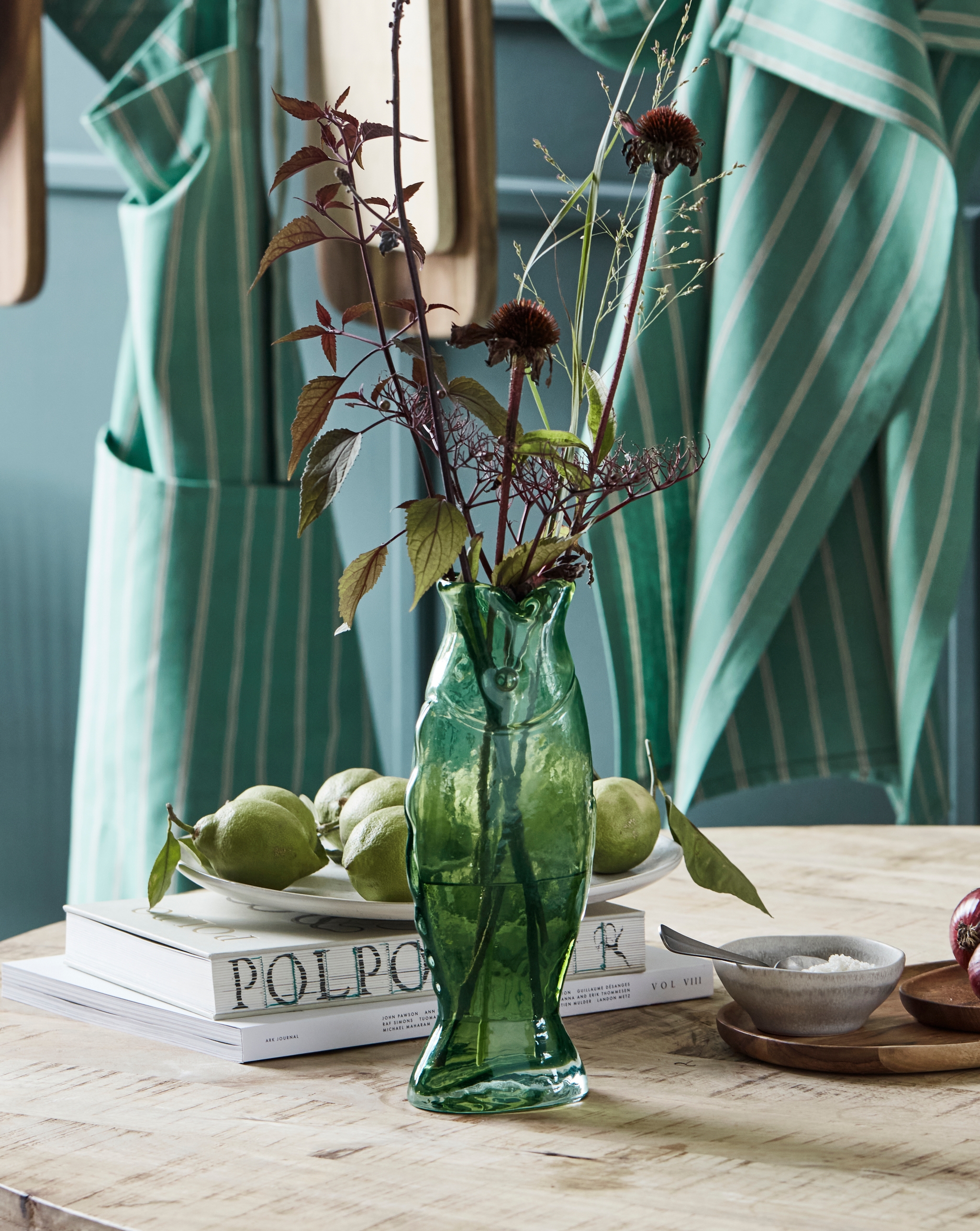 A green fish vase holding dark foliage sits on a wooden table with fruit, books, and small bowls, against a background of hanging striped aqua towels.